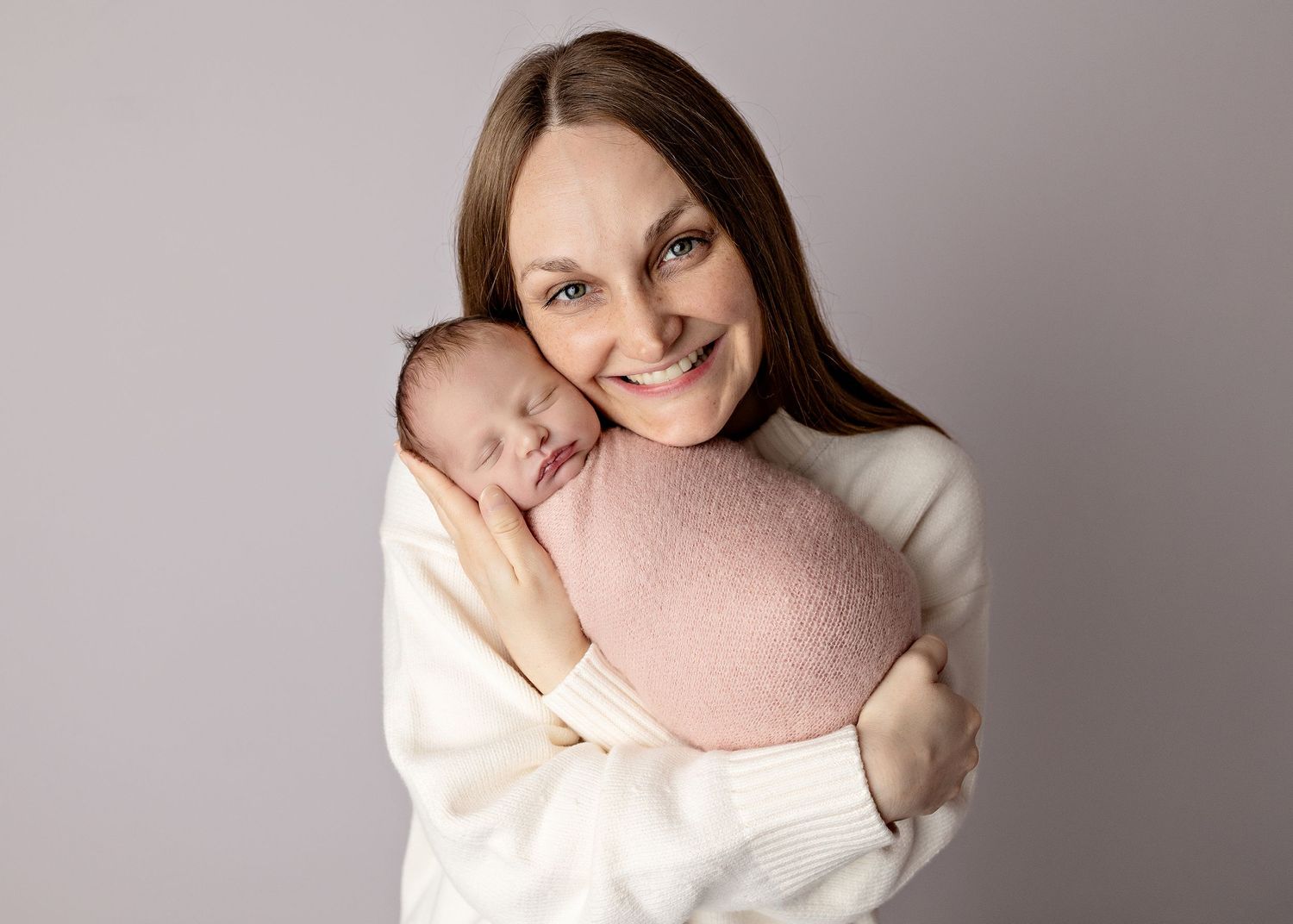 Smiling mother in white sweater holding her sleeping newborn baby wrapped in a pink blanket.