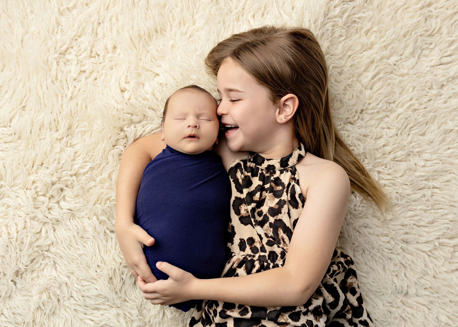 A smiling girl in leopard print dress holds a sleeping newborn wrapped in navy blue on a fluffy cream rug.