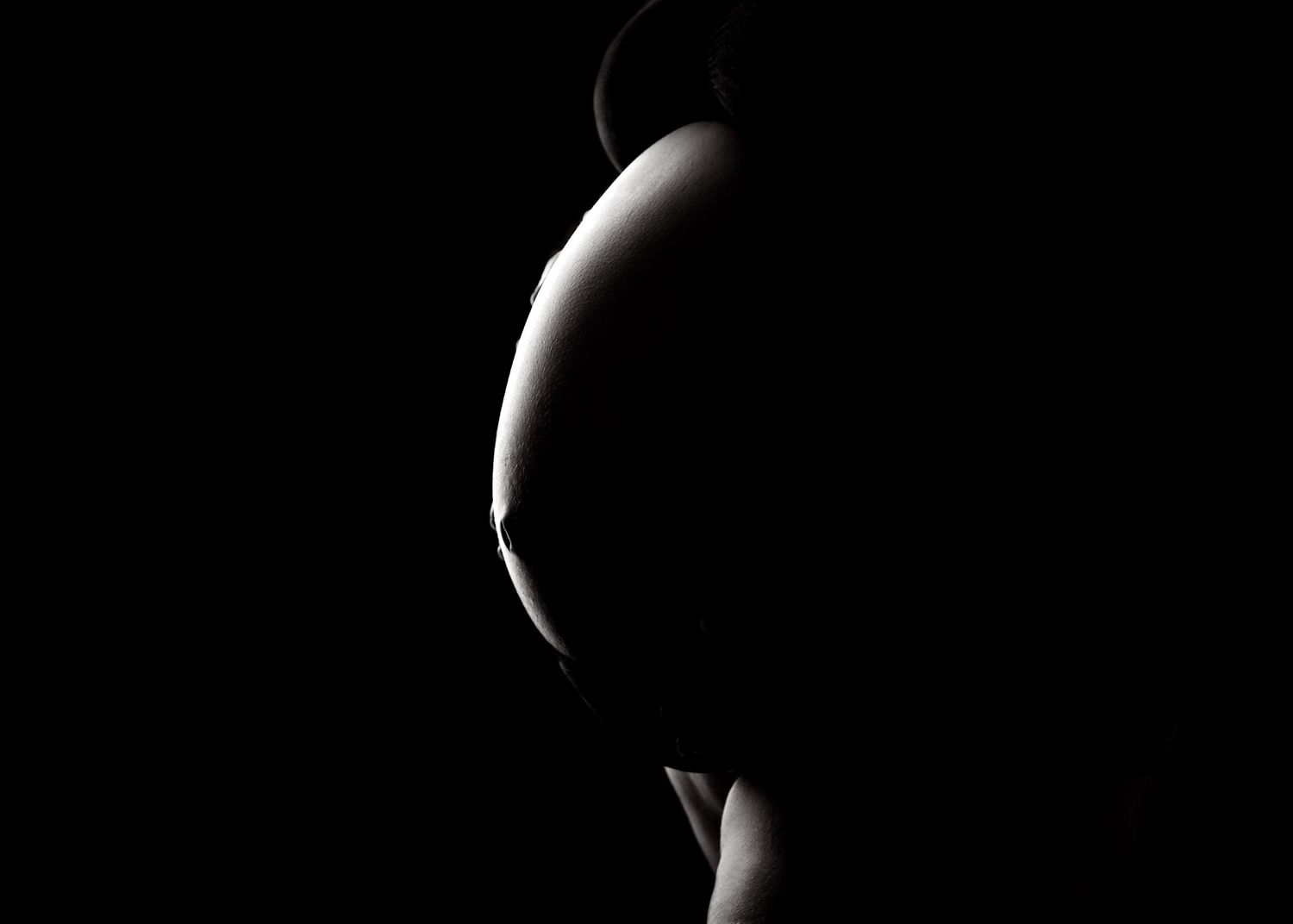 Silhouette of a pregnant woman's baby bump against a dark background in dramatic black and white maternity photography.