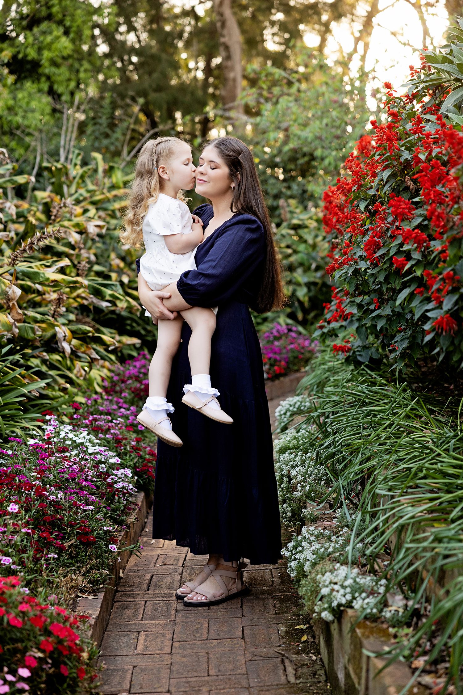 Tender family moments captured in a garden setting with red flowers and stone pathway.Toowoomba Photographer Toowoomba Newborn Photographer