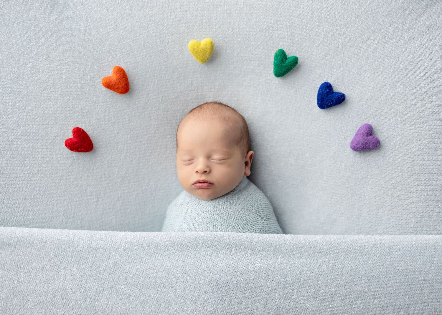 Sleeping newborn baby swaddled in blue wrap surrounded by colorful rainbow felt hearts on a soft light blue background.