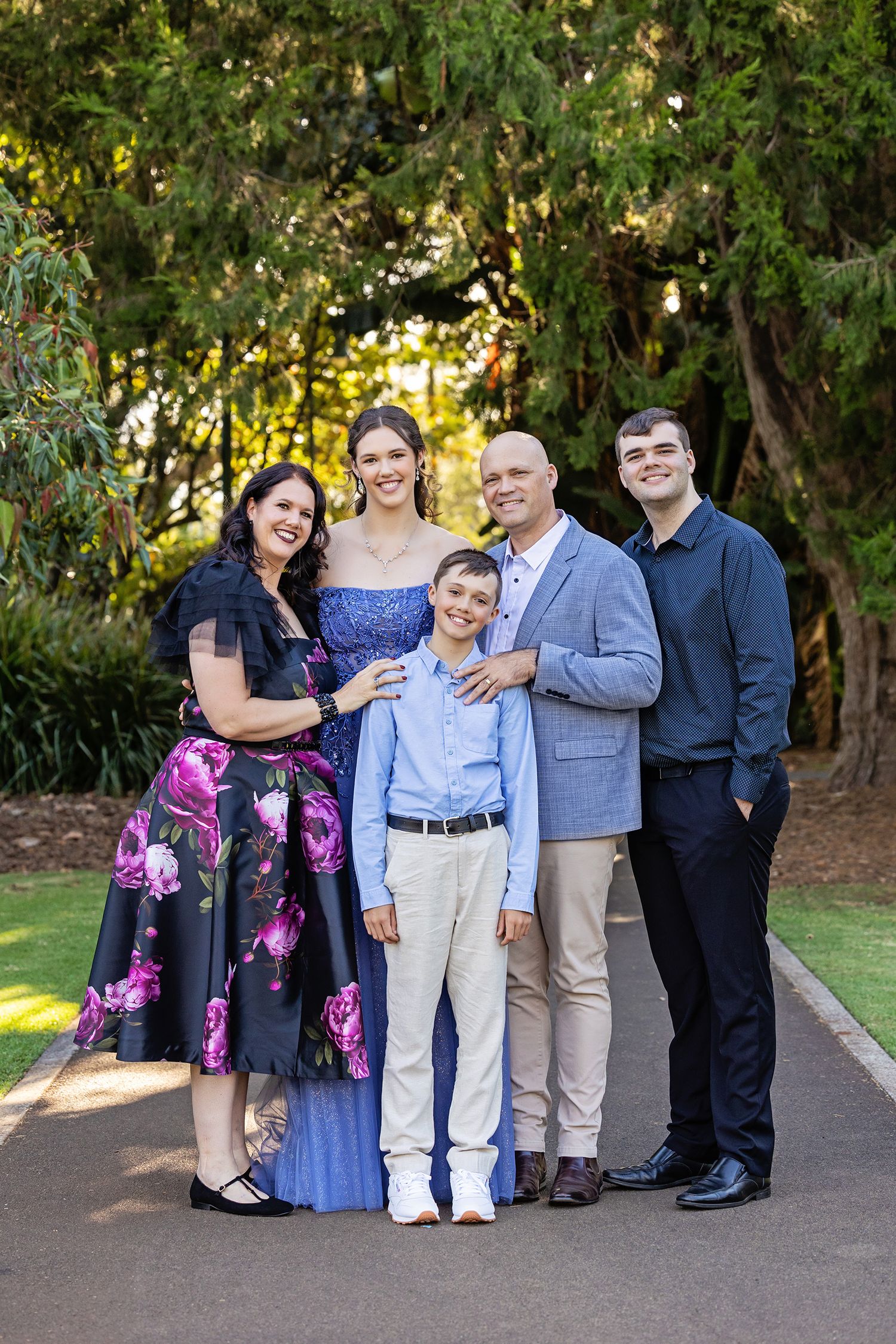 Family of five posing outdoors in formal attire, smiling together on a garden path surrounded by lush green trees.