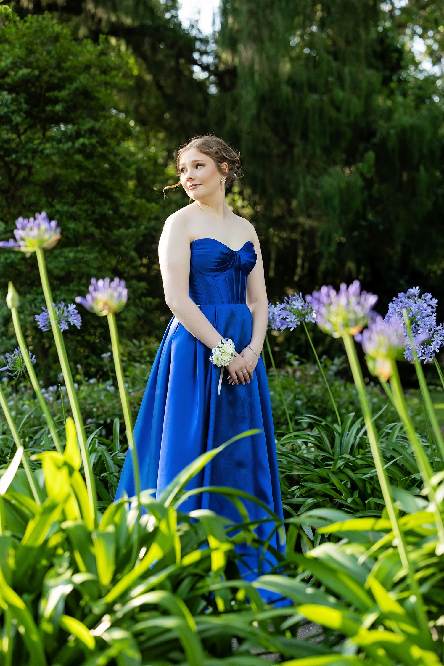 Young woman in royal blue strapless ball gown poses among purple agapanthus flowers in a lush garden.