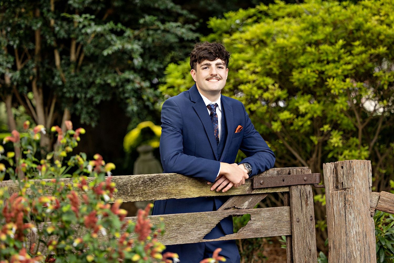 Formal portrait of person in navy suit leaning on rustic wooden fence in garden setting.Toowoomba Photographer Toowoomba Newborn Photographe