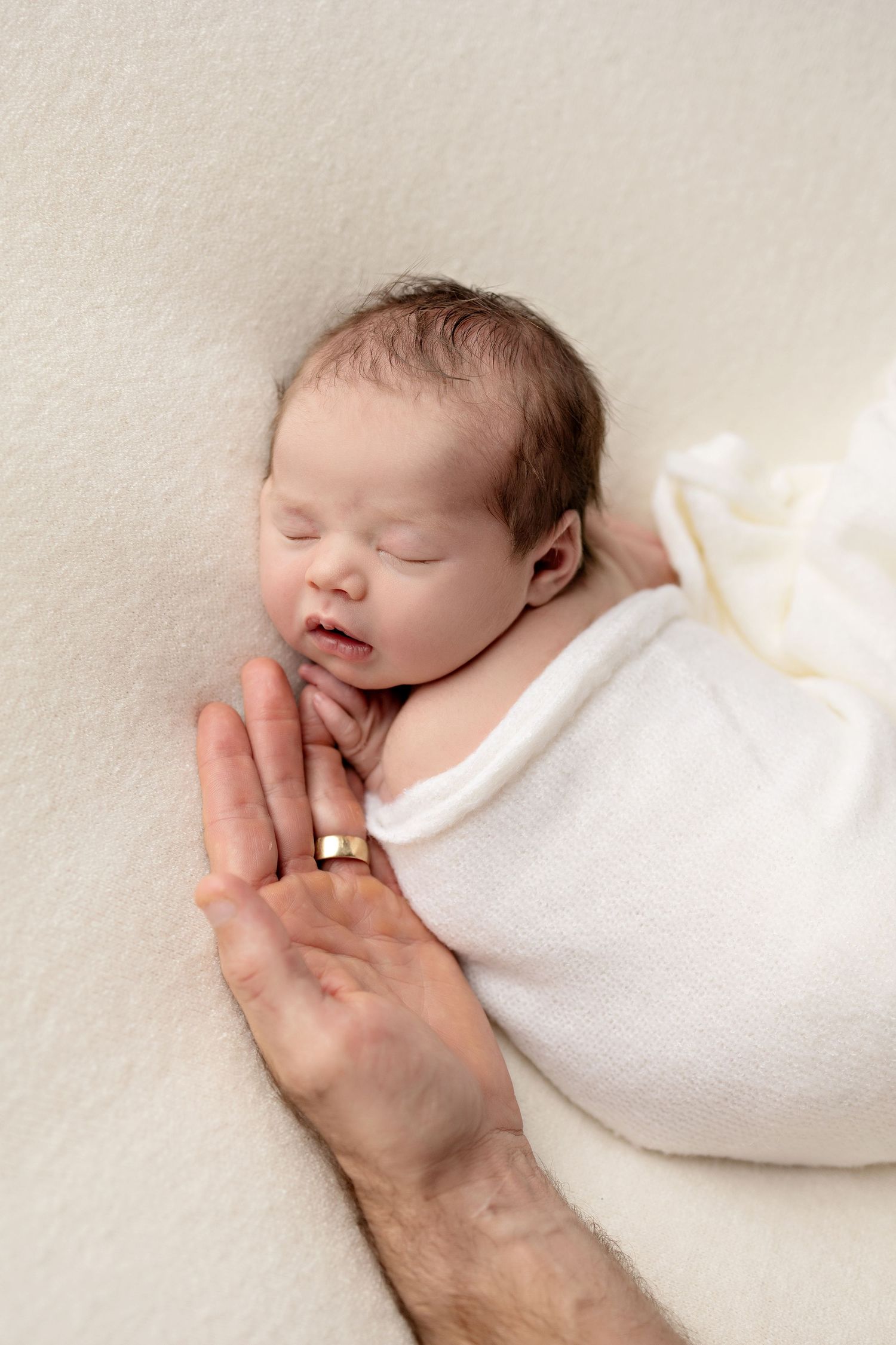 Sleeping newborn baby swaddled in white wrap with parent's hand wearing gold ring resting nearby on cream blanket.