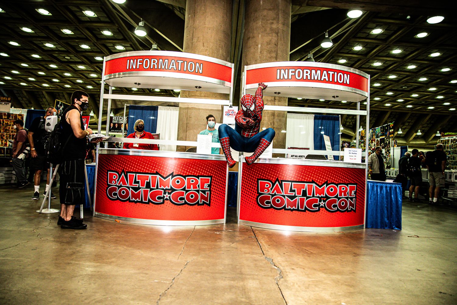 Spider-Man cosplayer posing at Baltimore Comic-Con information booth inside convention center.