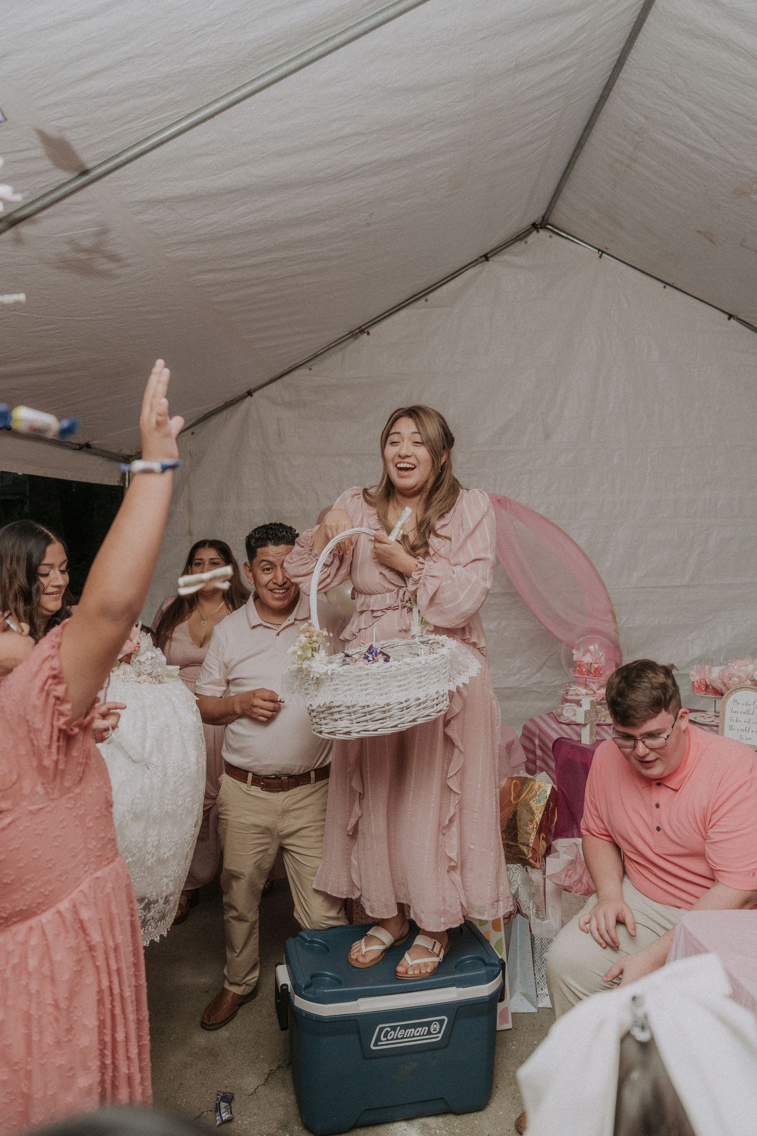 Baptism guests in pink dresses play a party game with a cooler at an outdoor tent reception.