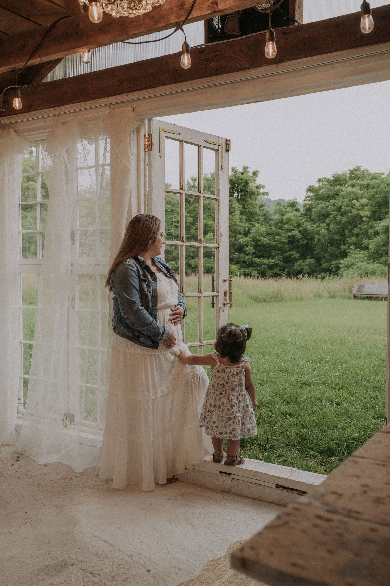 Mom and daughter stand in the doorway of a rustic glasshouse with string lights and white curtains.