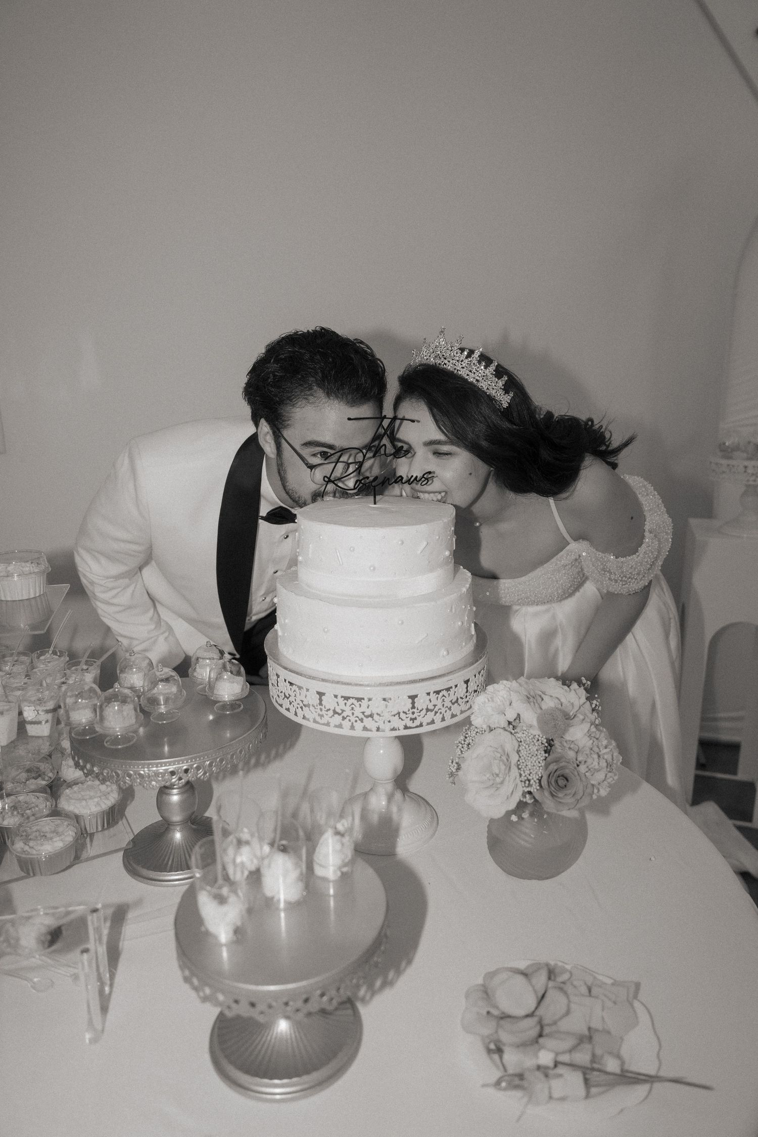 Black and white photo of a bride and groom kissing over a tiered wedding cake at a dessert table.