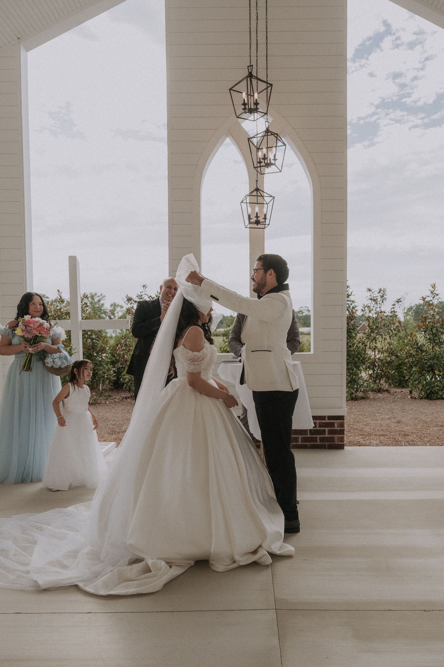 Bride and groom share first dance under elegant arched chapel with chandelier, bridesmaids in blue watching nearby.
