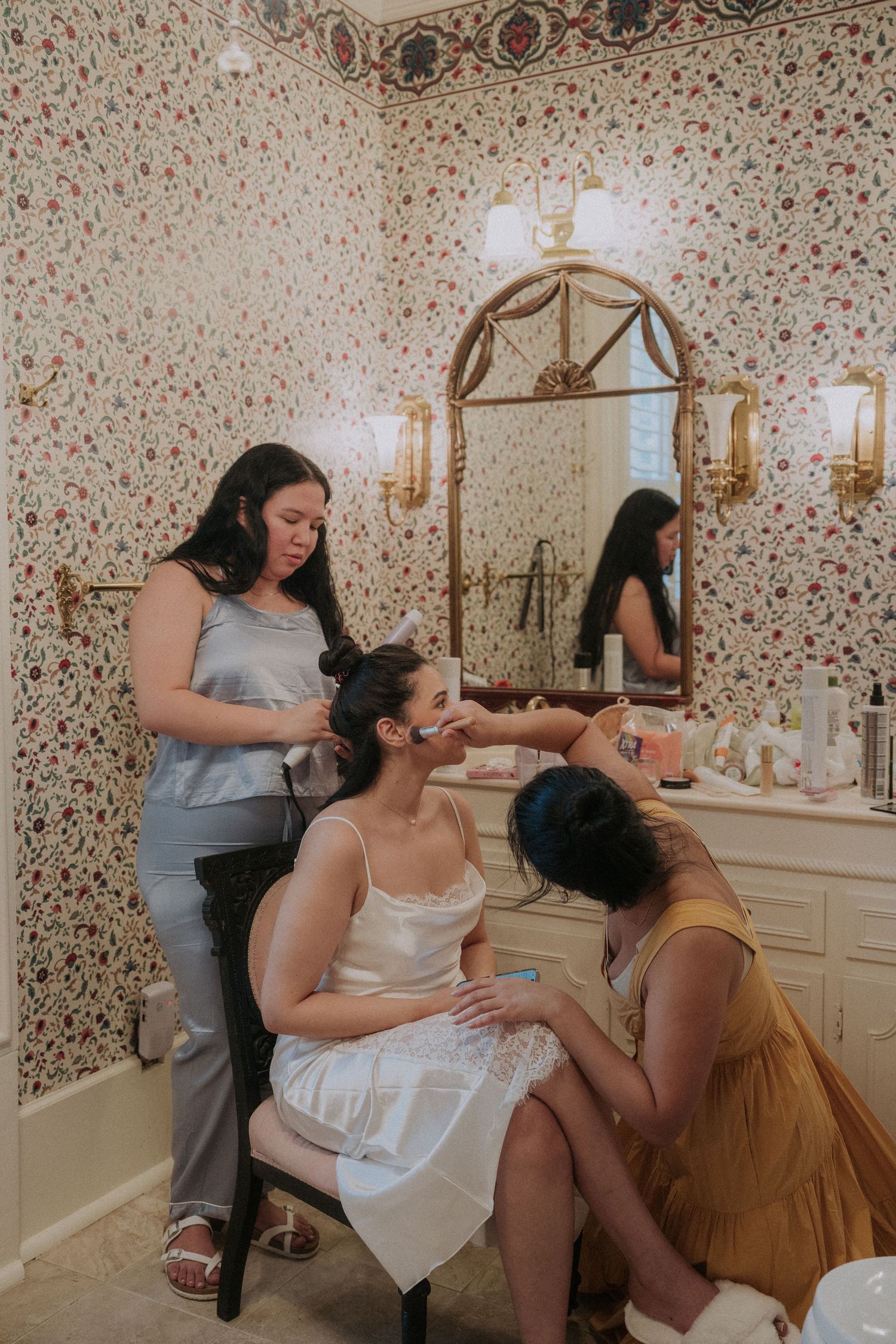 Bride getting hair and makeup done by two stylists in an elegant floral wallpapered dressing room with gold mirror.