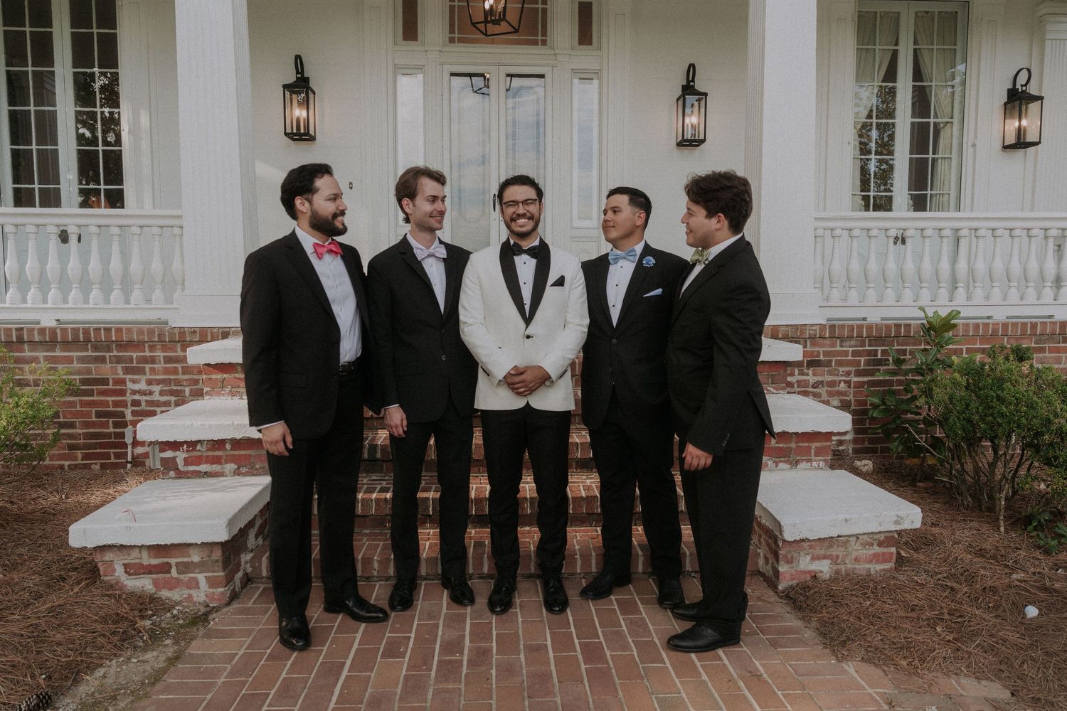 Groom in white tuxedo jacket stands with four groomsmen in black suits on brick steps outside elegant white venue.