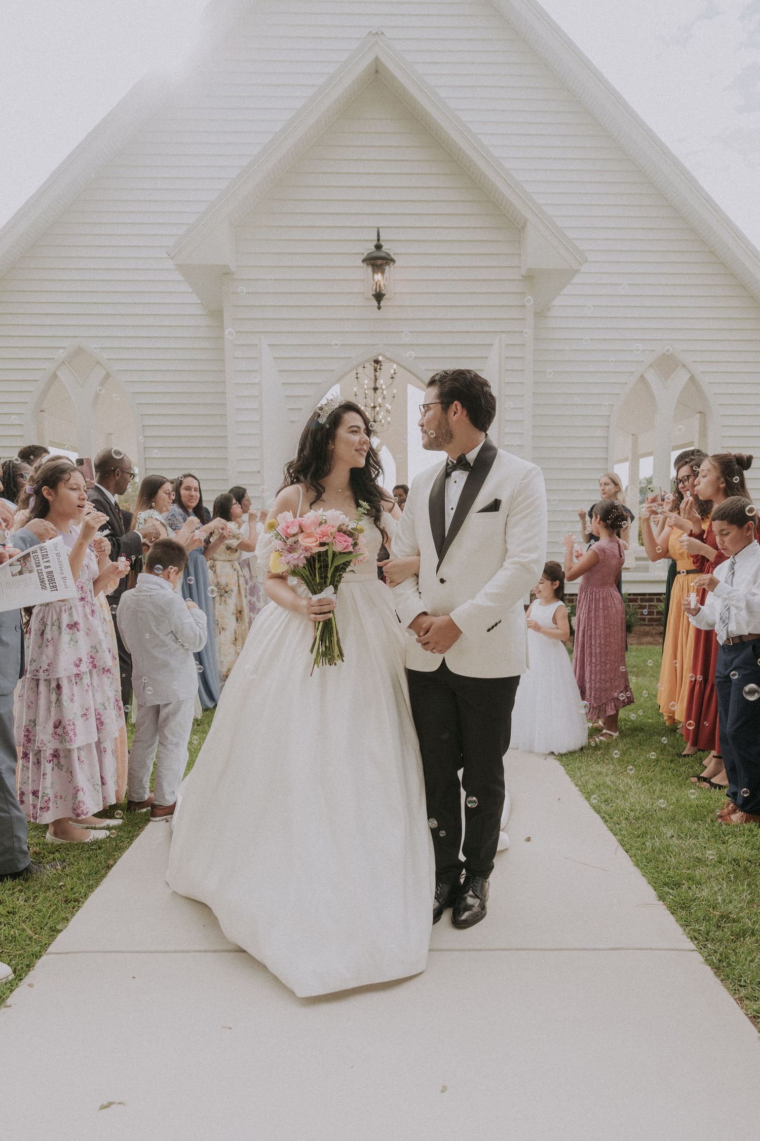Bride and groom walk down chapel path after wedding ceremony, guests cheering, bride holding pink bouquet.