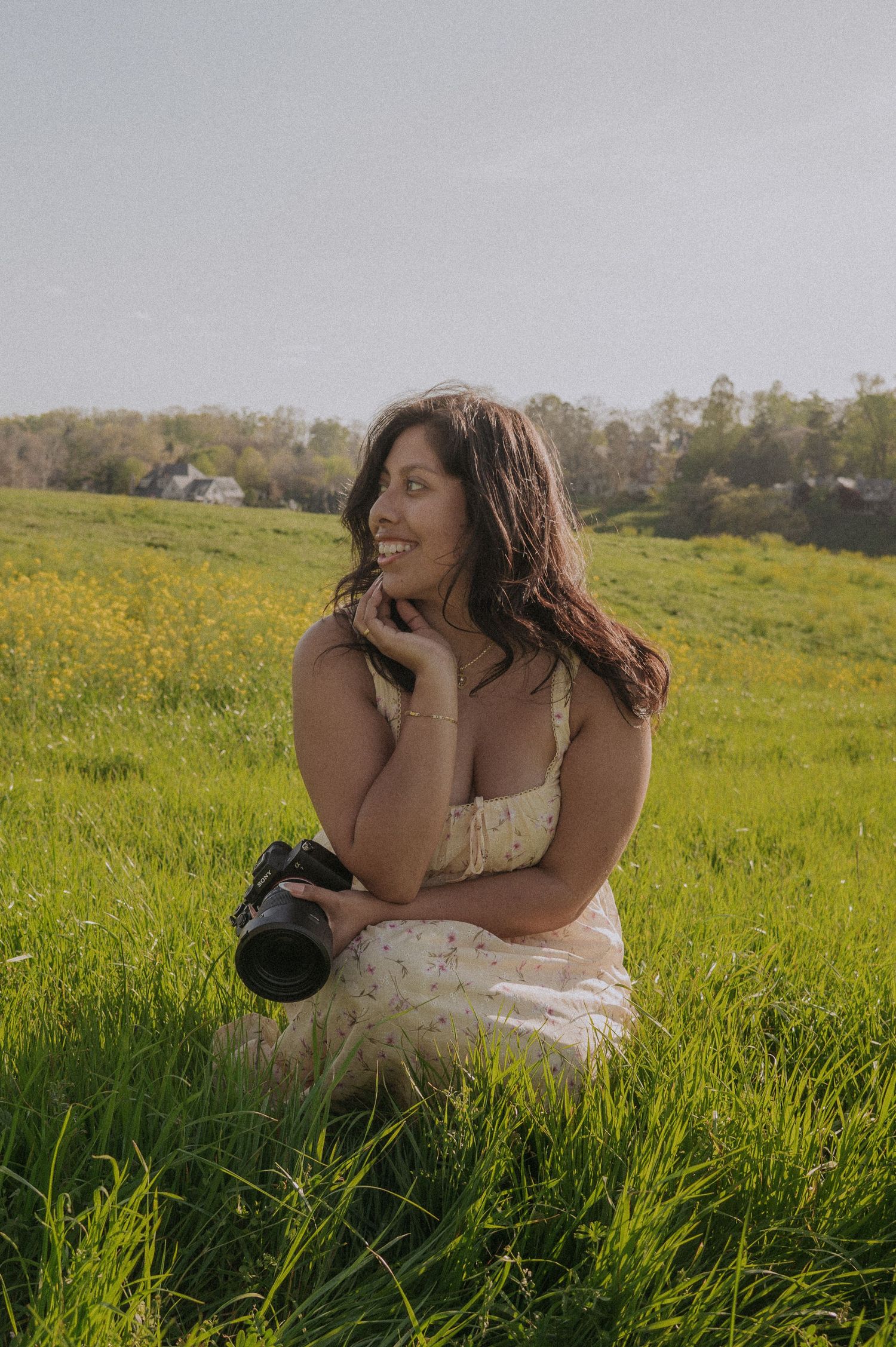 A smiling woman in a floral dress sits in a green meadow holding a camera, enjoying a sunny day outdoors.