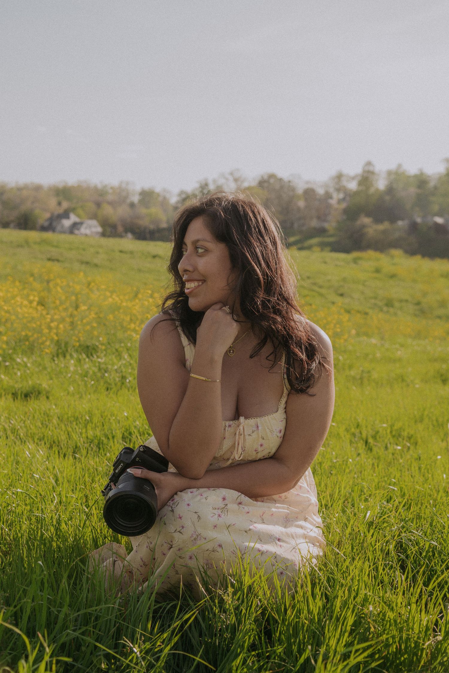A smiling woman in a floral dress sits in a green meadow holding a camera on a sunny day.