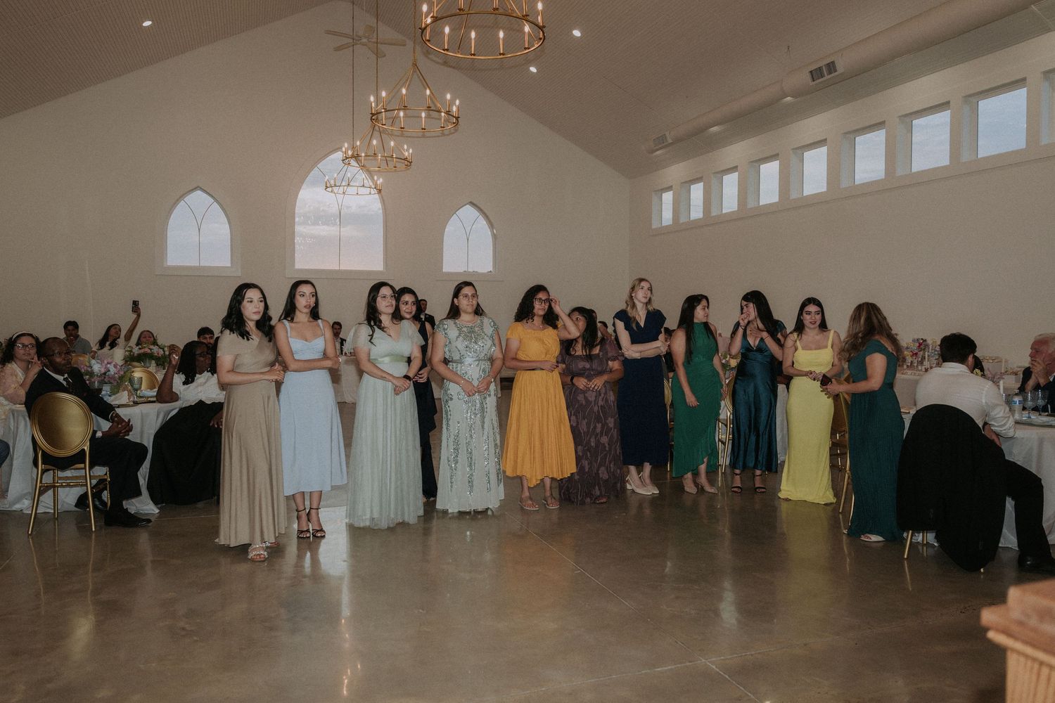 Bridesmaids in colorful mismatched dresses line up in an elegant white wedding reception venue with chandeliers.
