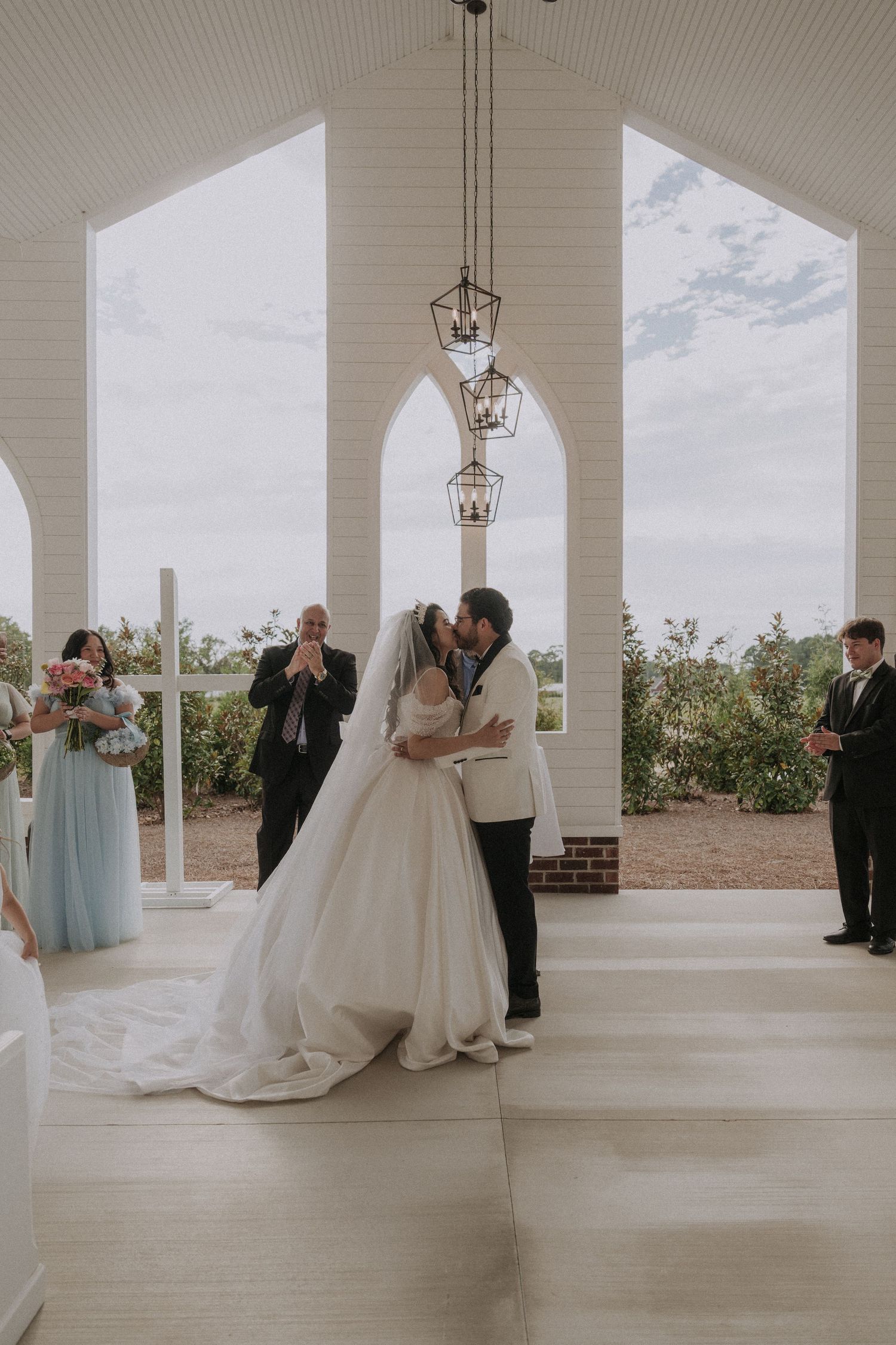 Bride and groom share first kiss at elegant white chapel wedding venue with arched windows and hanging lanterns.