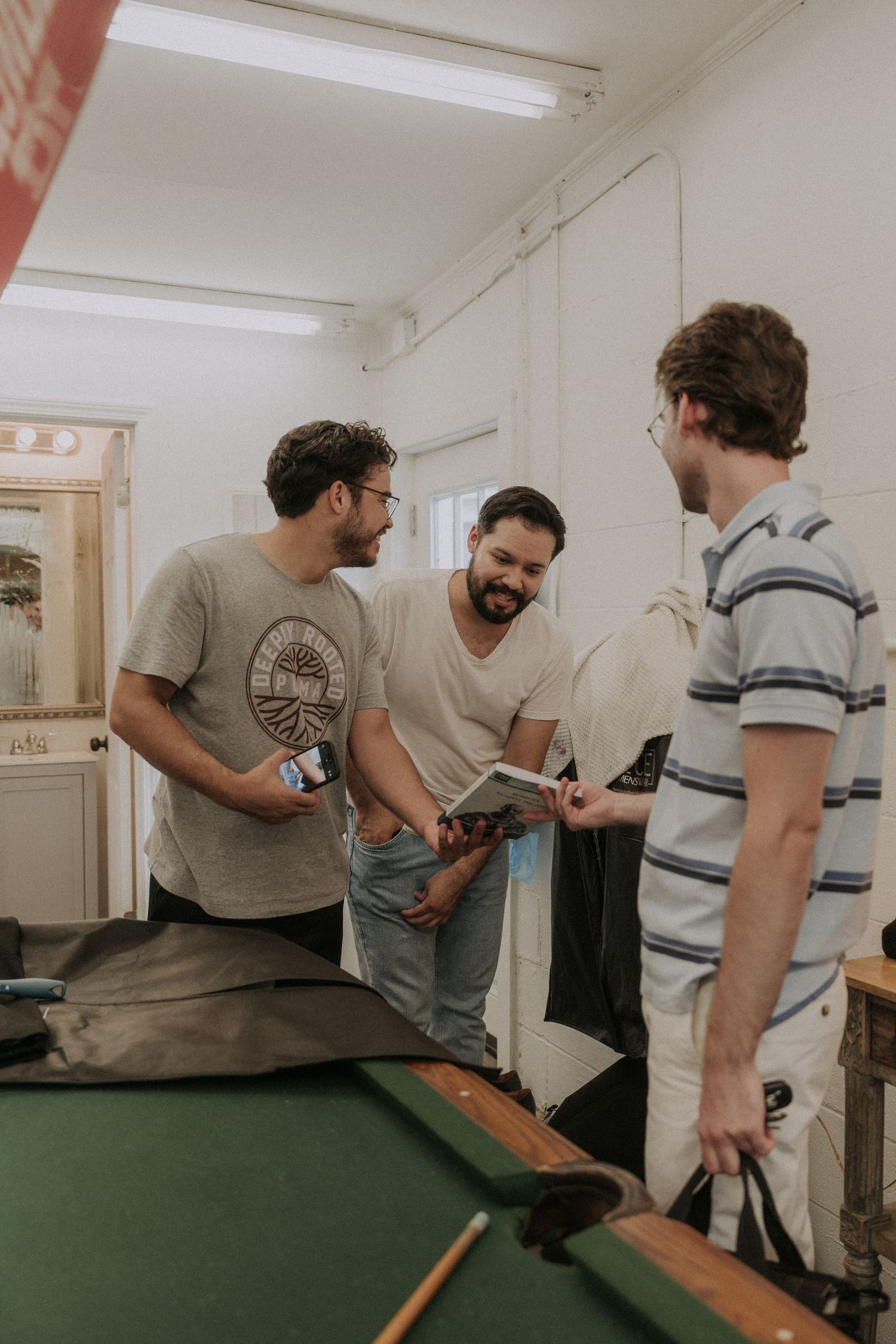 Three men socializing near a pool table in a casual indoor setting, sharing a book and smiling.