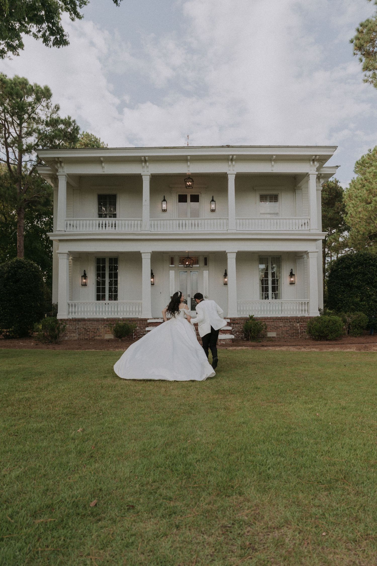 Bride and groom dancing on green lawn in front of elegant white two-story plantation-style wedding venue.