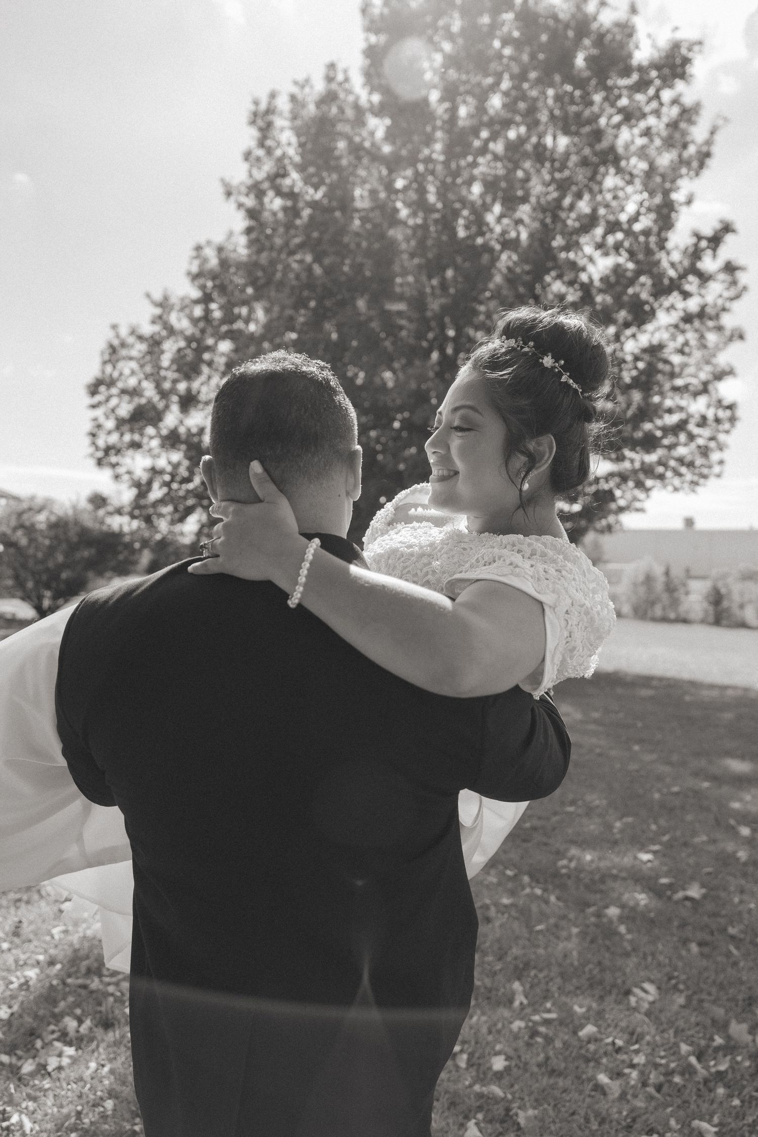 Black and white photo of a bride being carried by her groom in an outdoor setting.