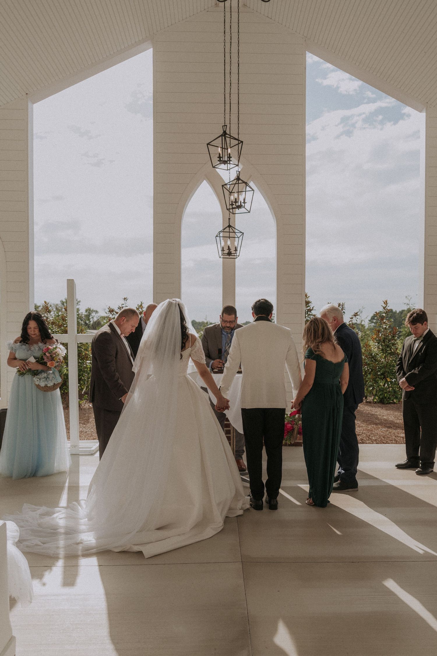 Bride in white gown walks down chapel aisle with wedding party during outdoor ceremony with arched windows.