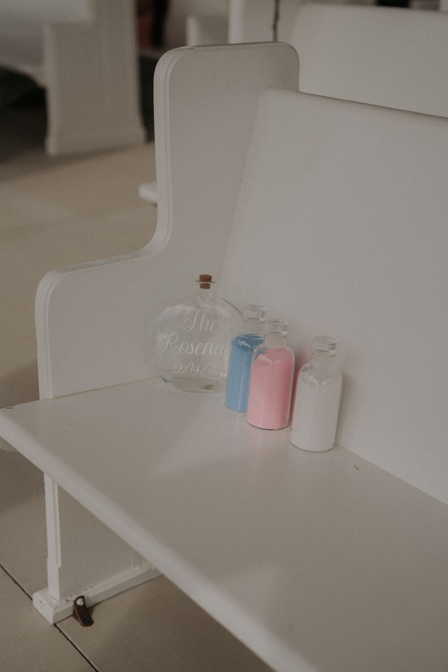 White church pew with sand ceremony bottles in pink, blue, and white beside a glass unity vase.