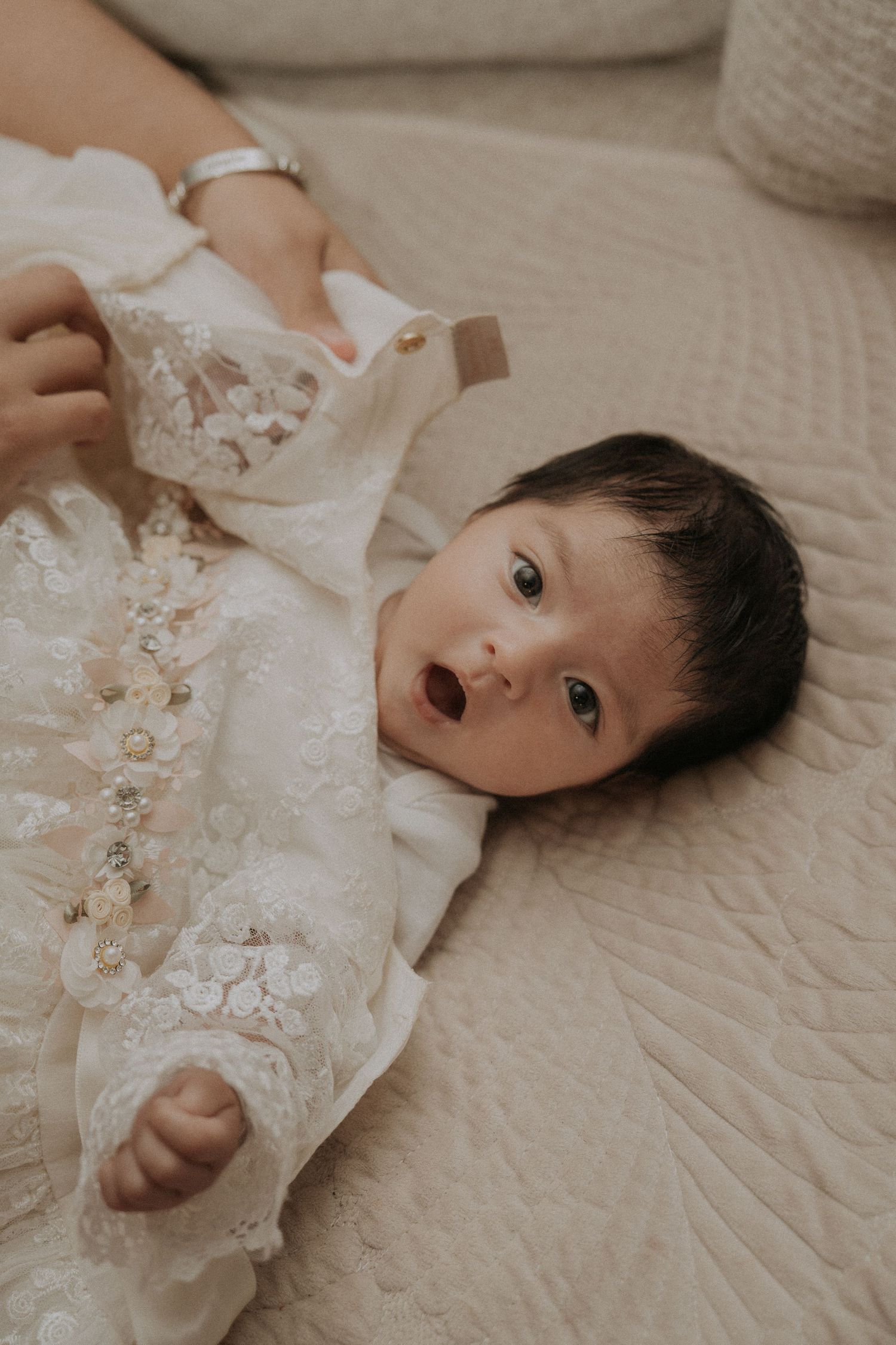 Baby wearing white lace outfit lies on quilted bedding while smiling.