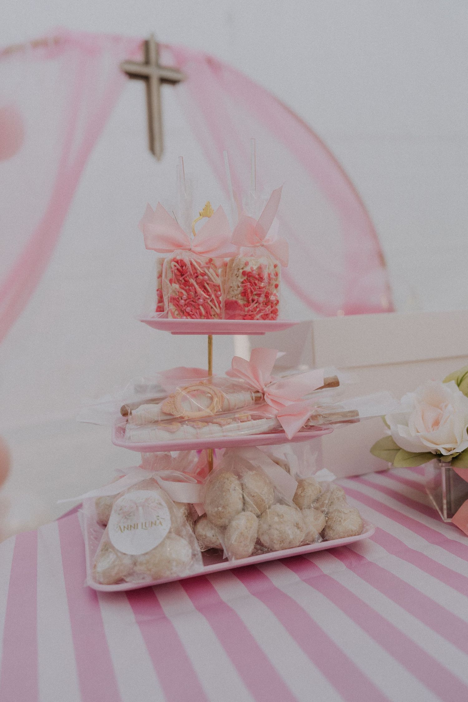 Pink and white tiered dessert display with cross decoration on striped tablecloth.