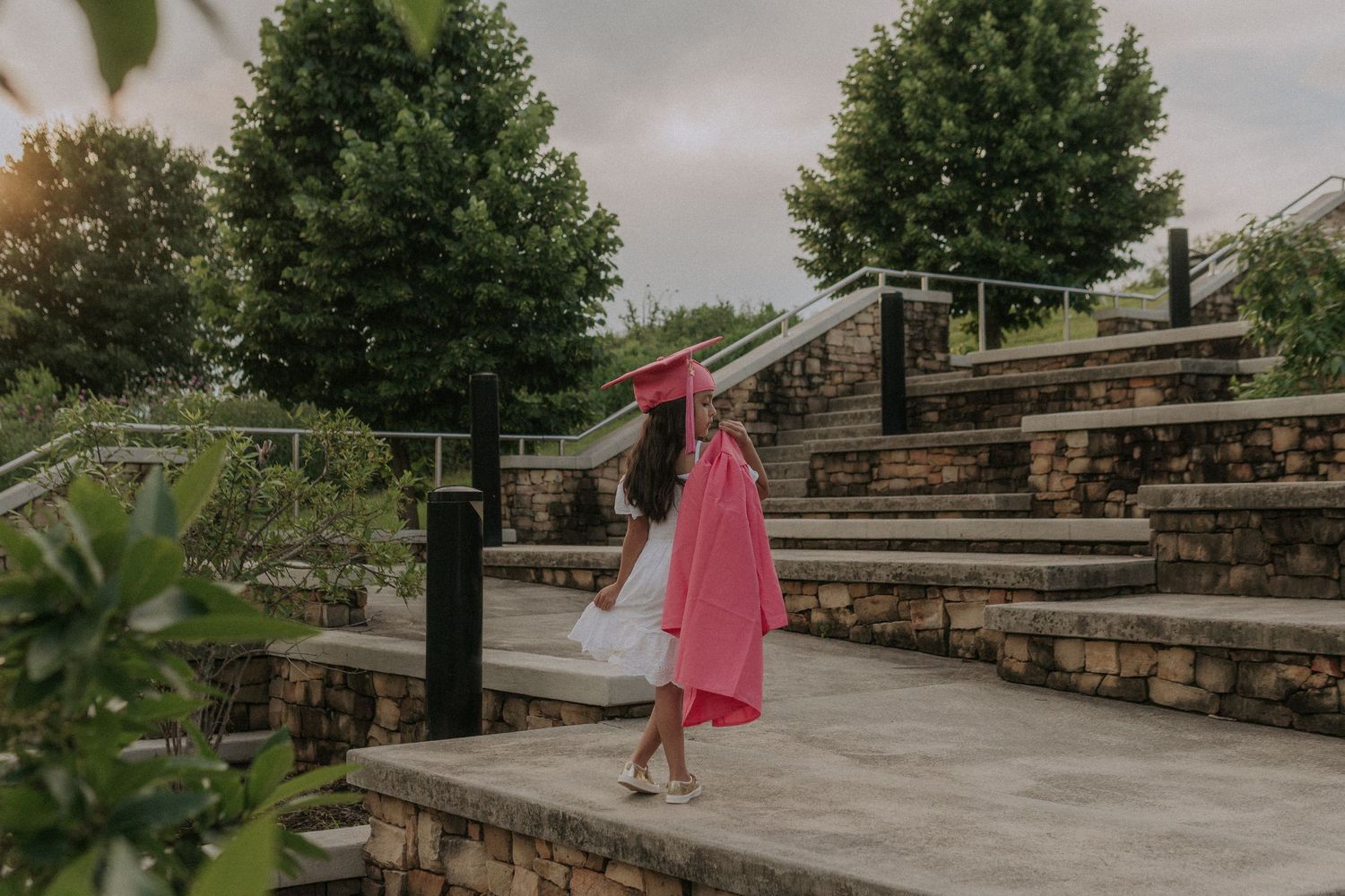 A girl stands on a winding stone pathway in a pink graduation cap and gown thleads through a landscaped garden with lush greenery and trees.