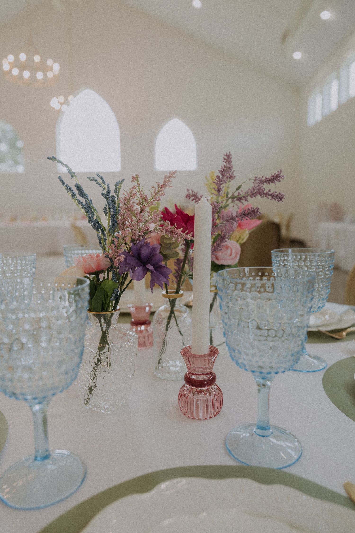Elegant wedding table setting with blue hobnail glasses, pink candle holders, and wildflower centerpieces in a bright venue.