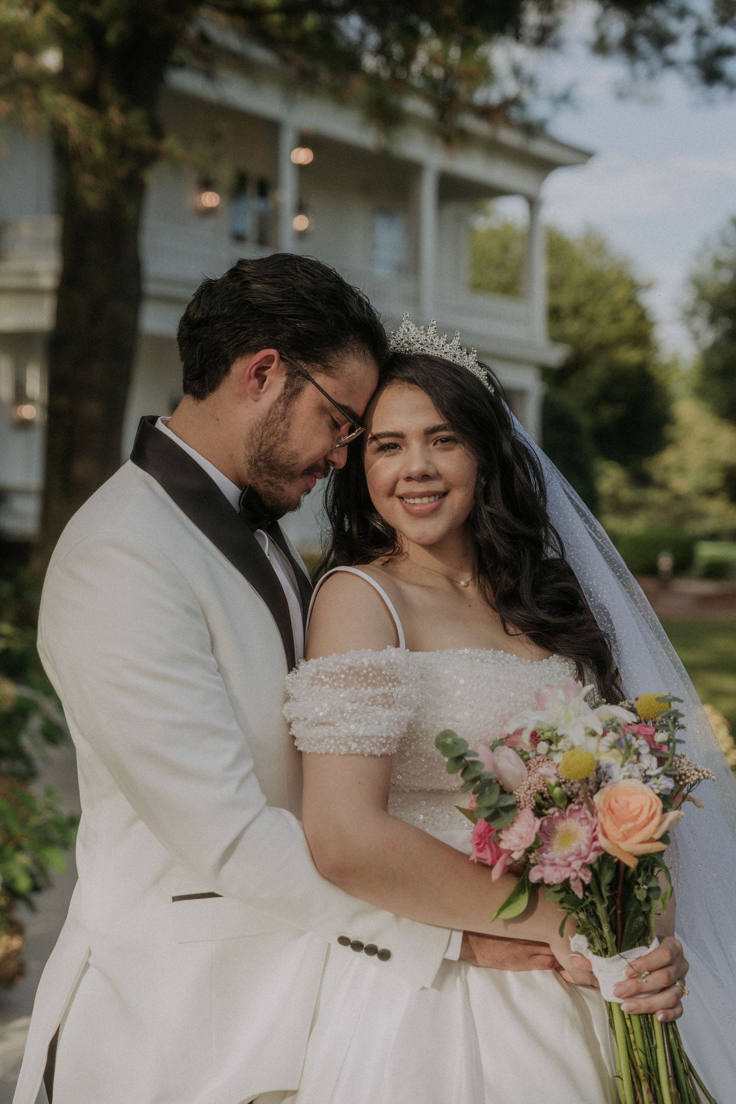 A bride in a white gown and tiara holds a bouquet while her groom in a white tuxedo embraces her outdoors.