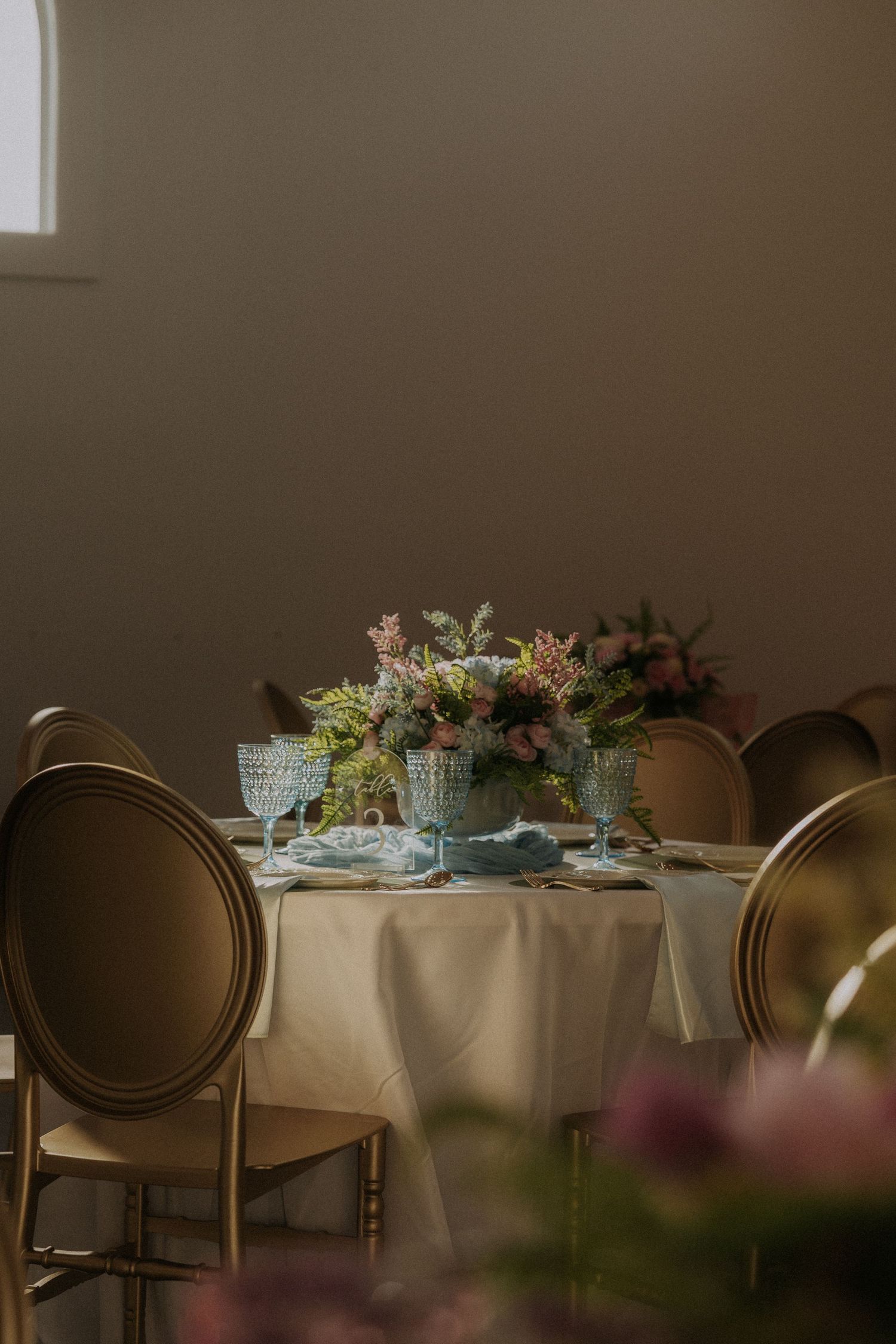 Elegant wedding table setting with colorful floral centerpiece and gold chairs in soft natural light.