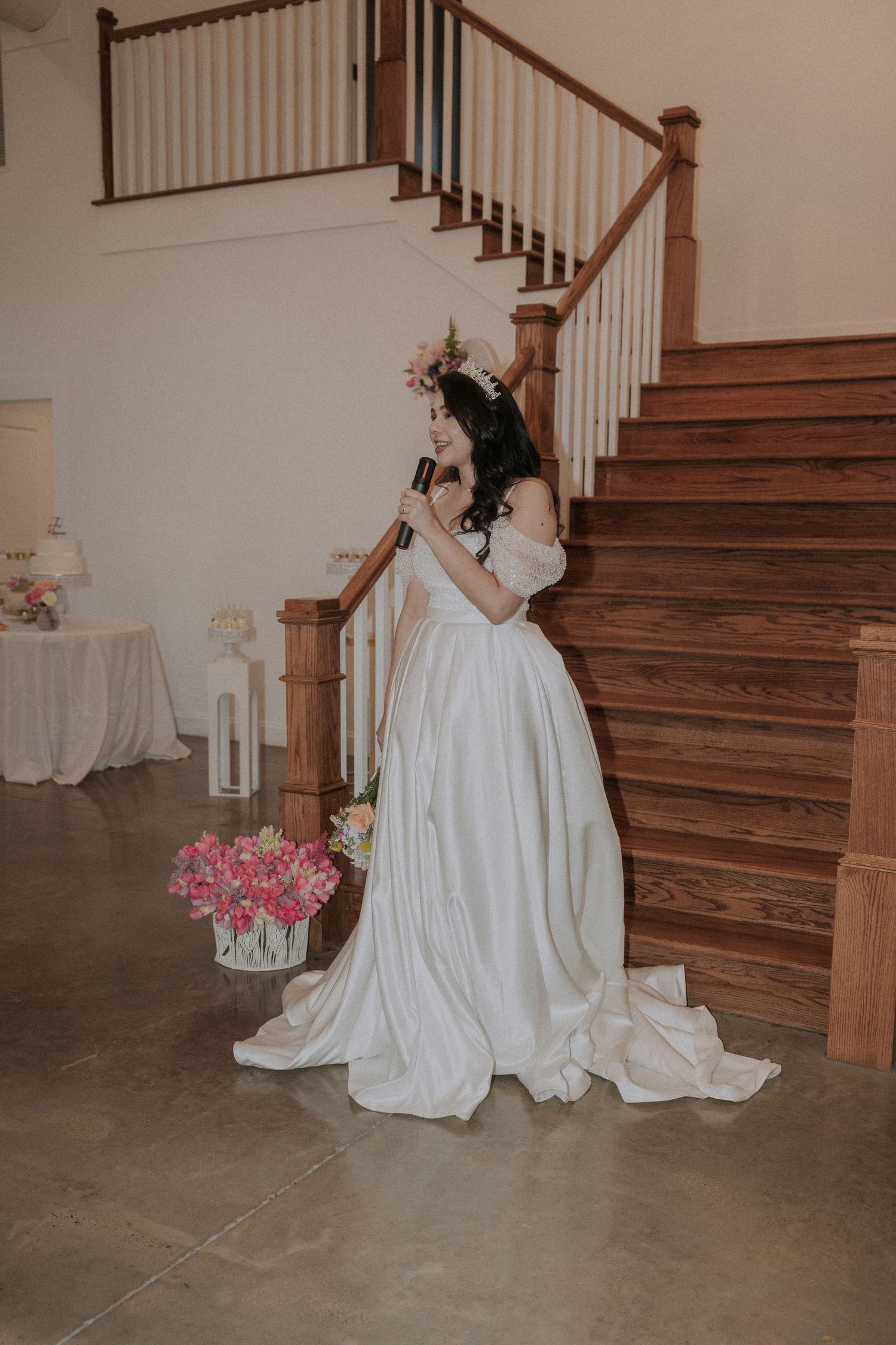 Bride in white off-shoulder gown wearing floral crown stands by wooden staircase holding microphone.