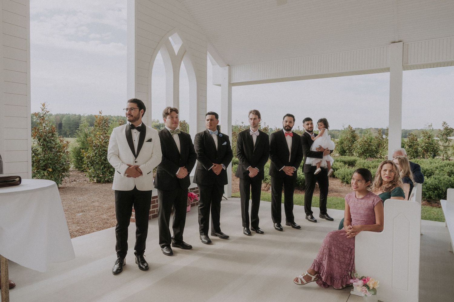 Groom in white tuxedo stands with groomsmen in black suits during outdoor wedding ceremony on white porch.