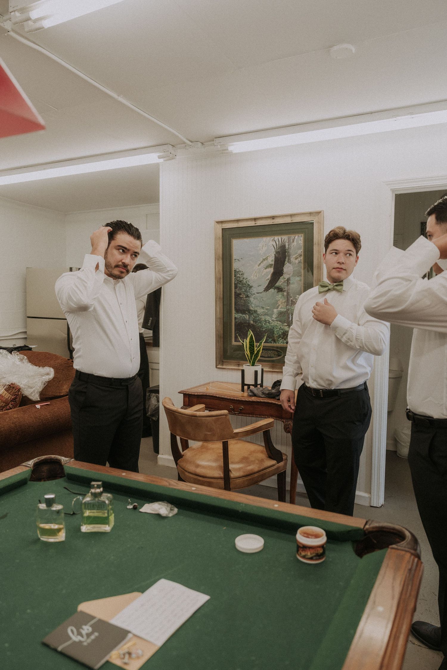 Three groomsmen in white shirts and black pants prepare for a wedding near a green pool table.