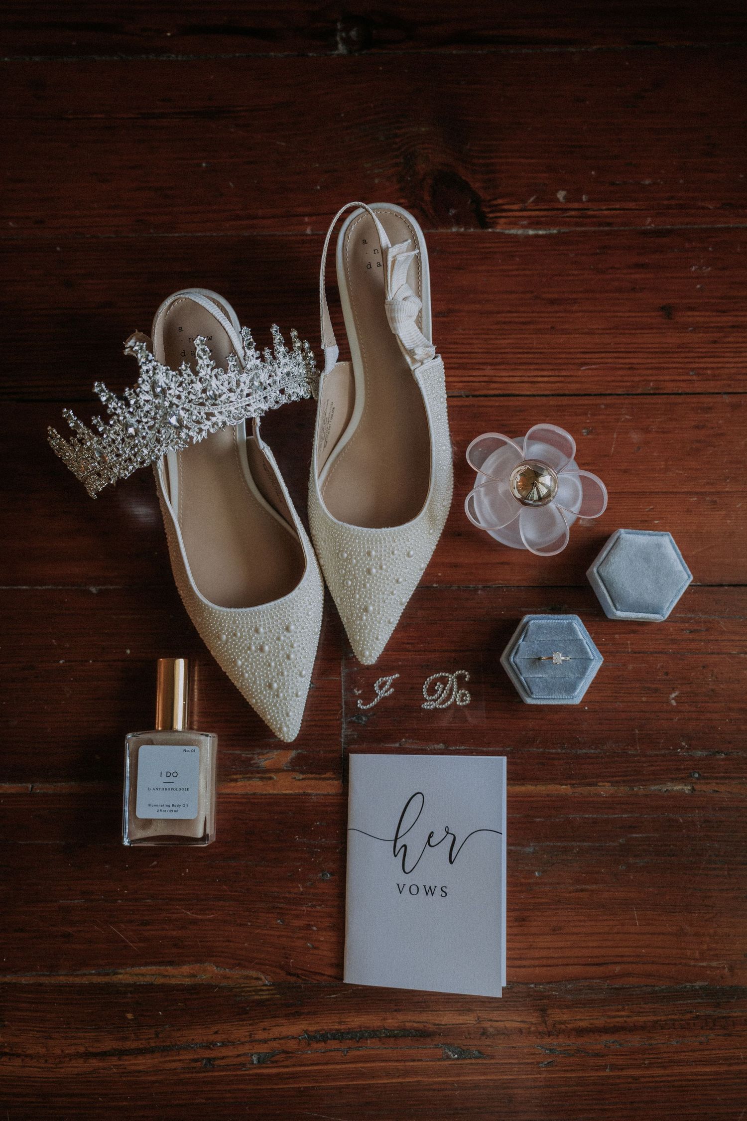 Bridal flat shoes with pearls, crystal tiara, vow book, ring box, and perfume arranged on rustic wood floor.