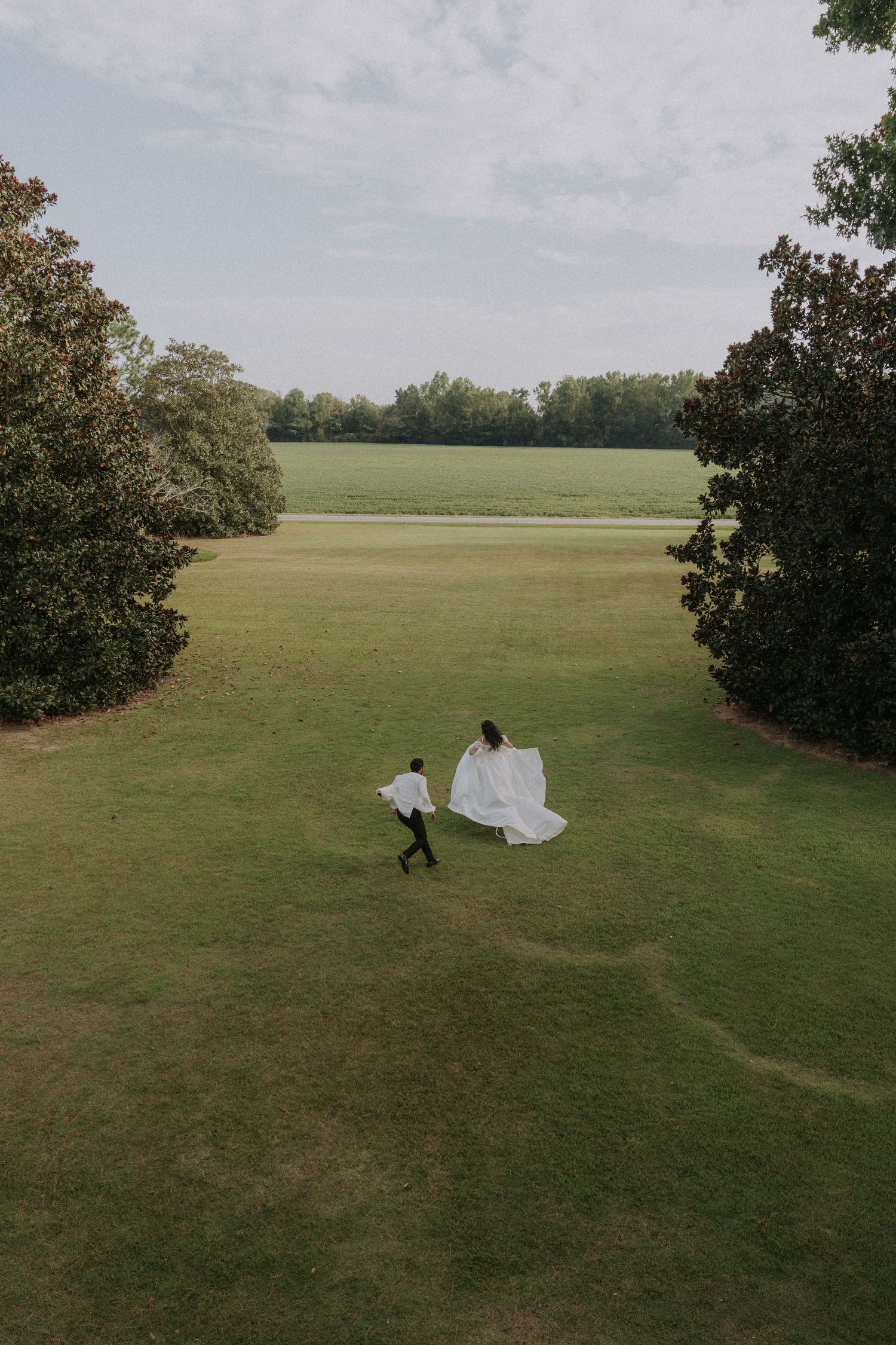 Aerial view of bride and groom running across lush green lawn surrounded by trees at outdoor wedding.