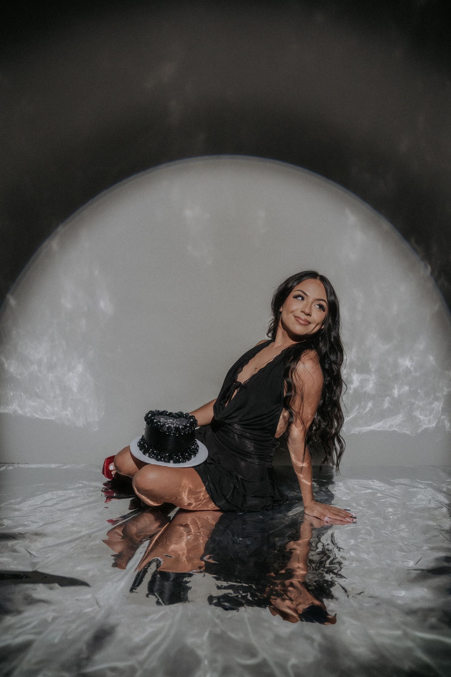Artistic portrait in black dress sitting in reflective water against dramatic white circular backdrop.