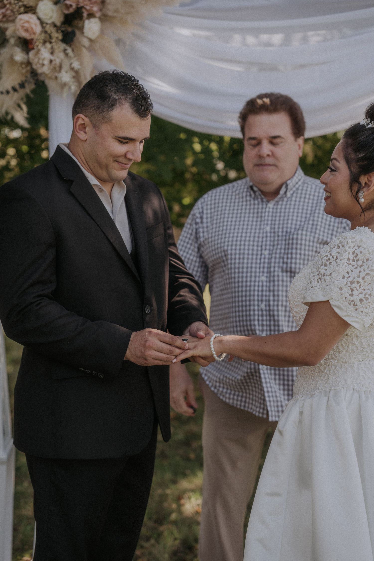 Exchange of rings during outdoor wedding ceremony under white fabric arch.