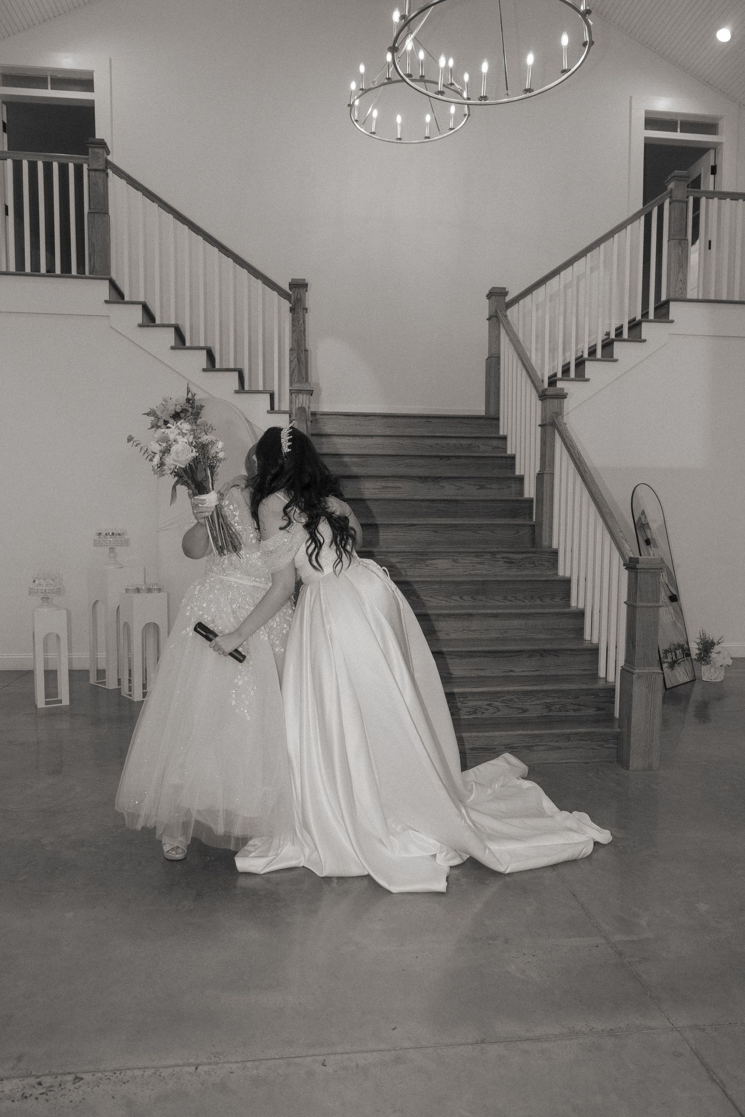 Black and white photo of bride in flowing white gown embracing young flower girl by grand staircase.