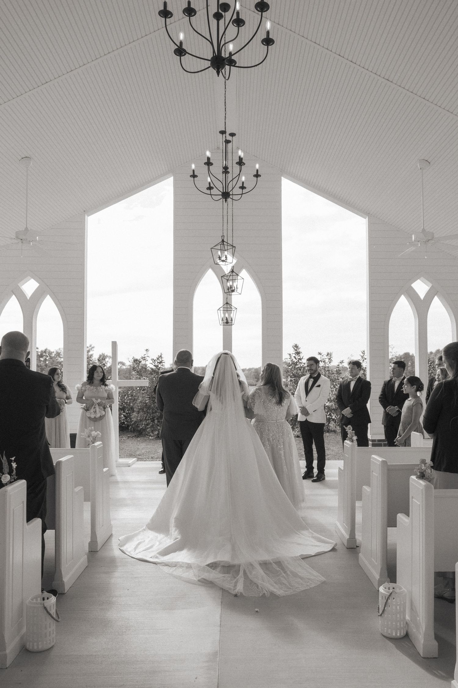 Black and white photo of bride walking down chapel aisle toward groom at altar.