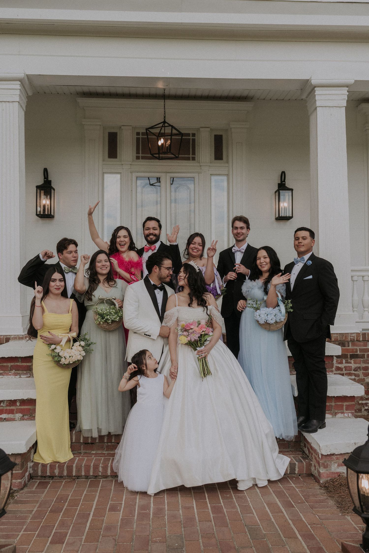 Wedding party posing on brick steps of elegant venue, bride in white gown holding bouquet with groom in white tuxedo.