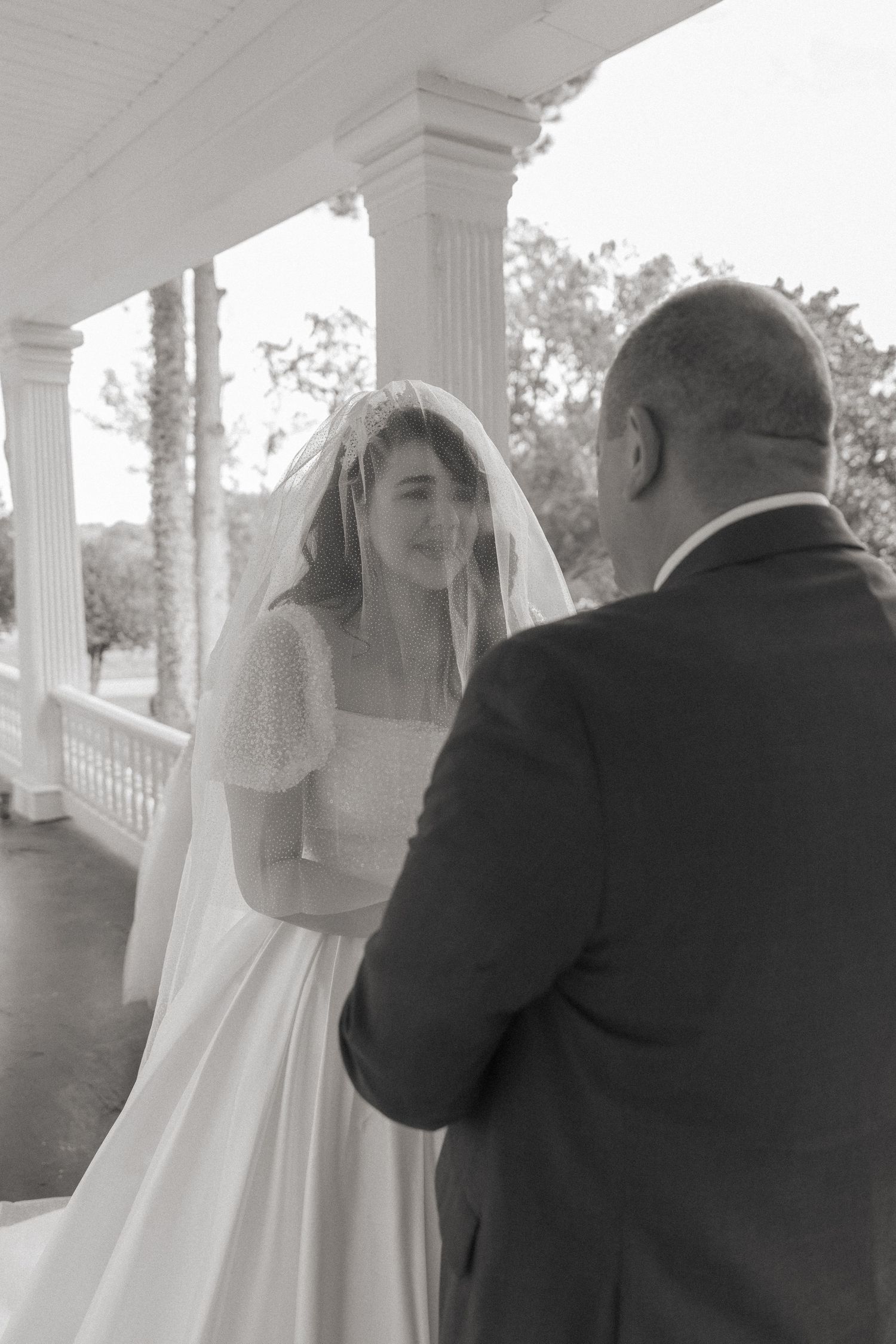 Black and white photo of a bride in a veil and gown facing a man in a suit on a classic white columned porch.