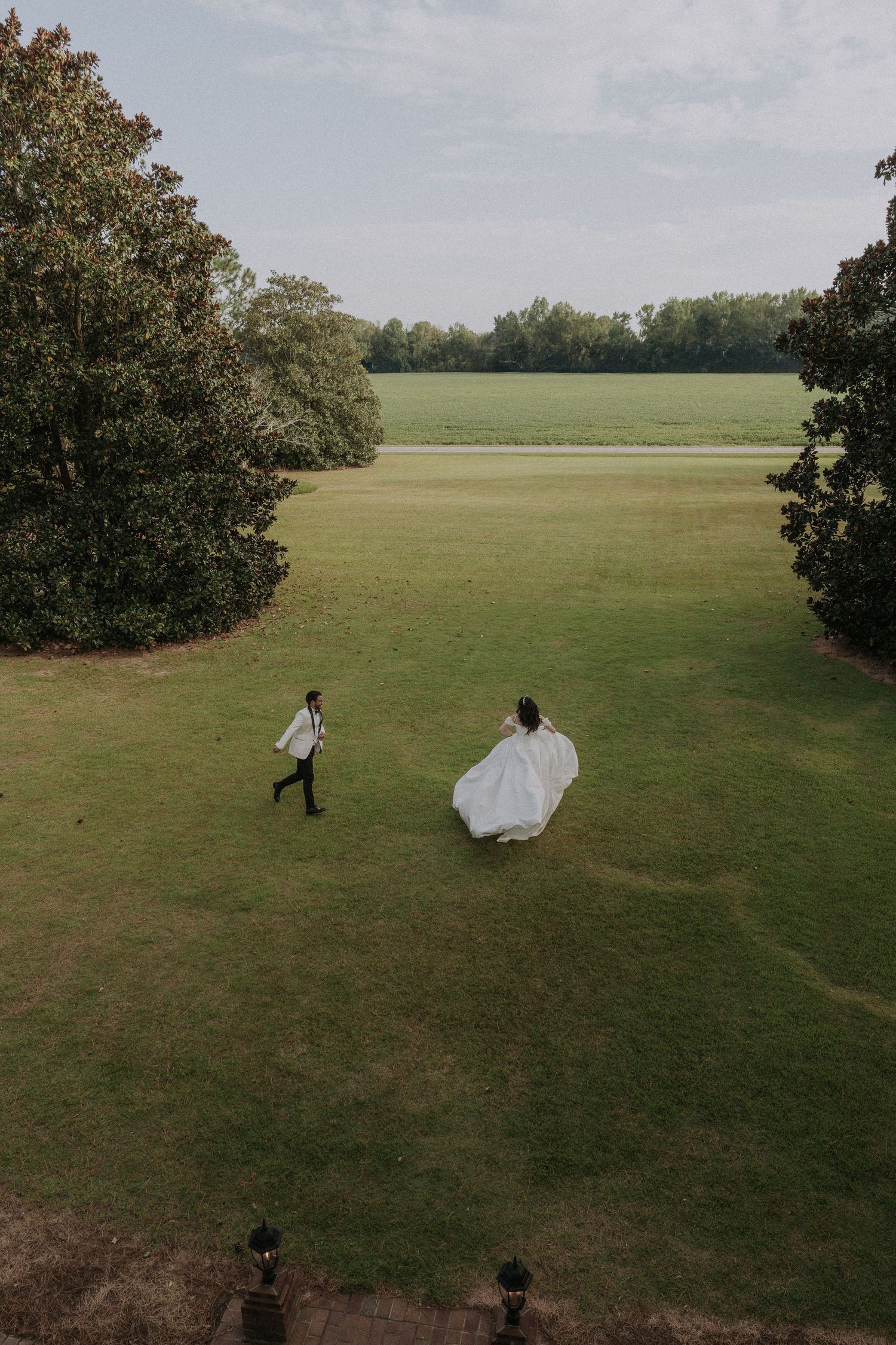 A bride and groom run playfully across a vast green field on a sunny day.