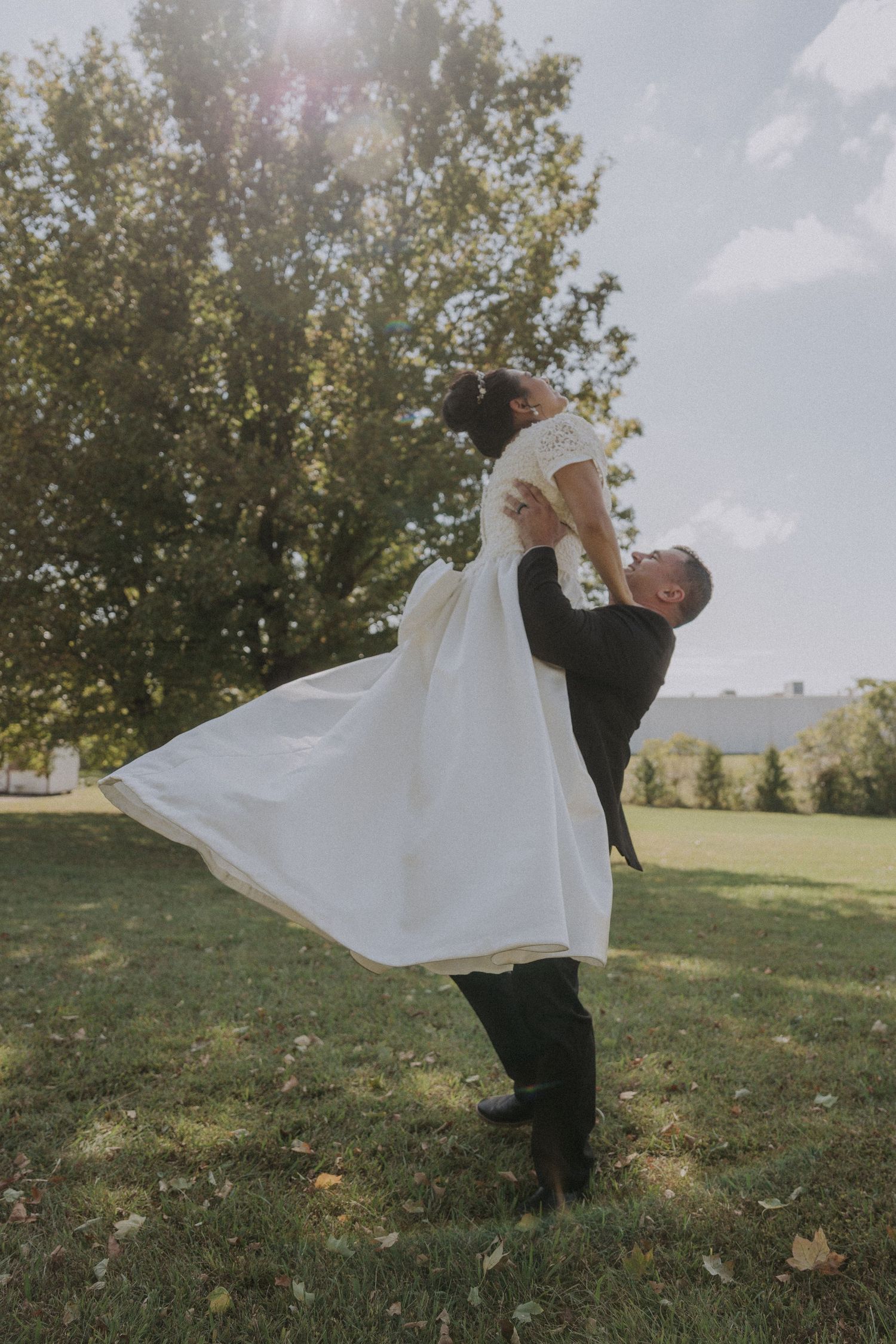 A romantic wedding moment captured outdoors as the groom lifts the bride in a flowing white dress against a sunlit backdrop.
