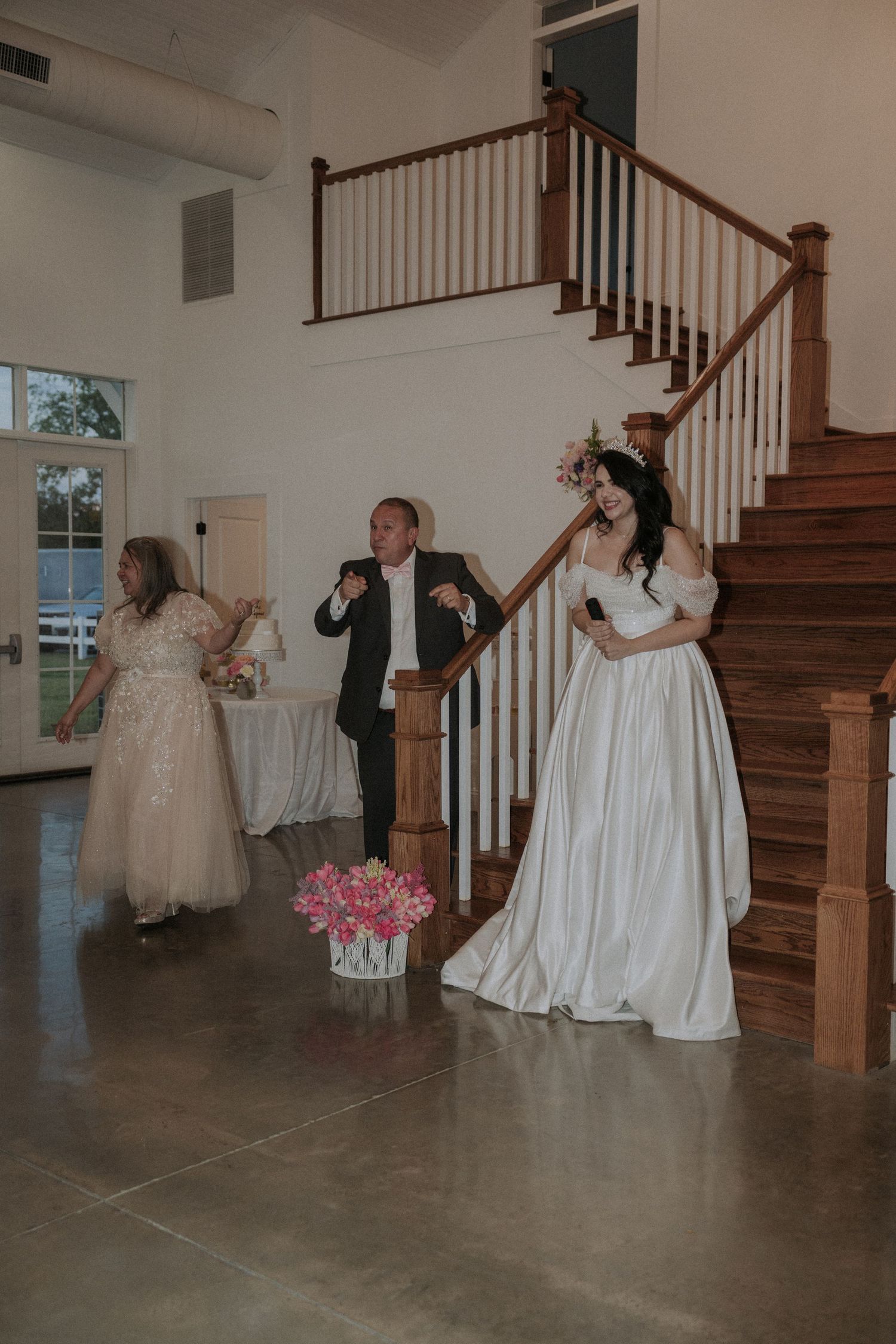 Bride in white gown stands near wooden staircase with guests during wedding reception in elegant venue.