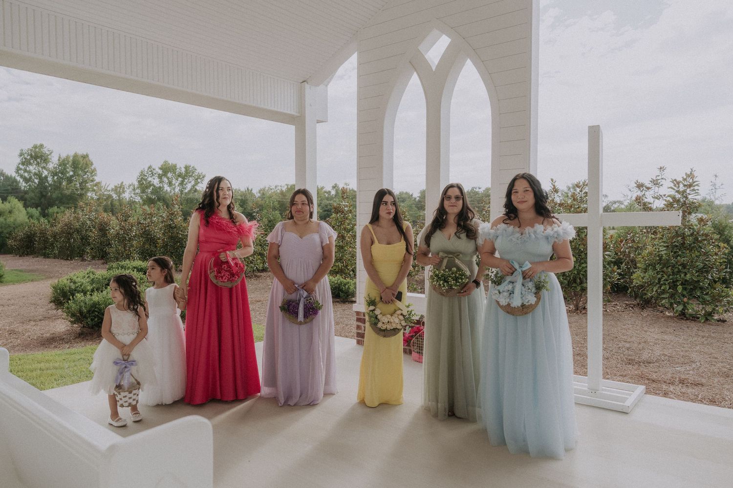 Bridesmaids in colorful dresses holding bouquets stand under white arched chapel with garden backdrop.