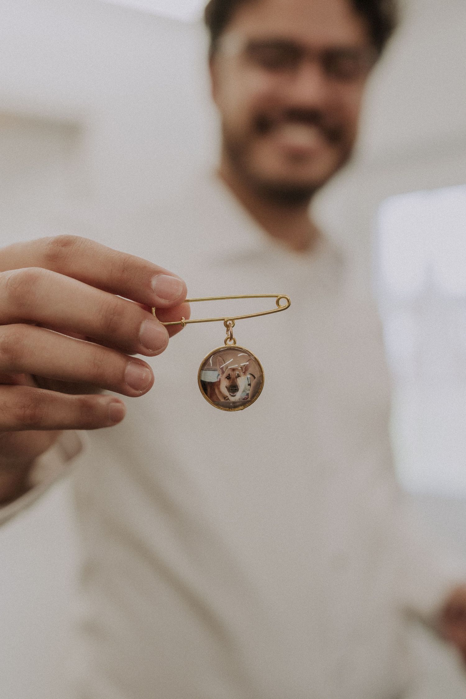Smiling man holding a gold safety pin brooch with a small round pet portrait charm in focus.