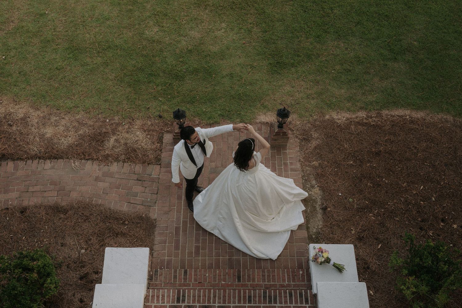 Aerial view of bride and groom dancing on brick pathway, white wedding dress flowing, surrounded by green lawn.