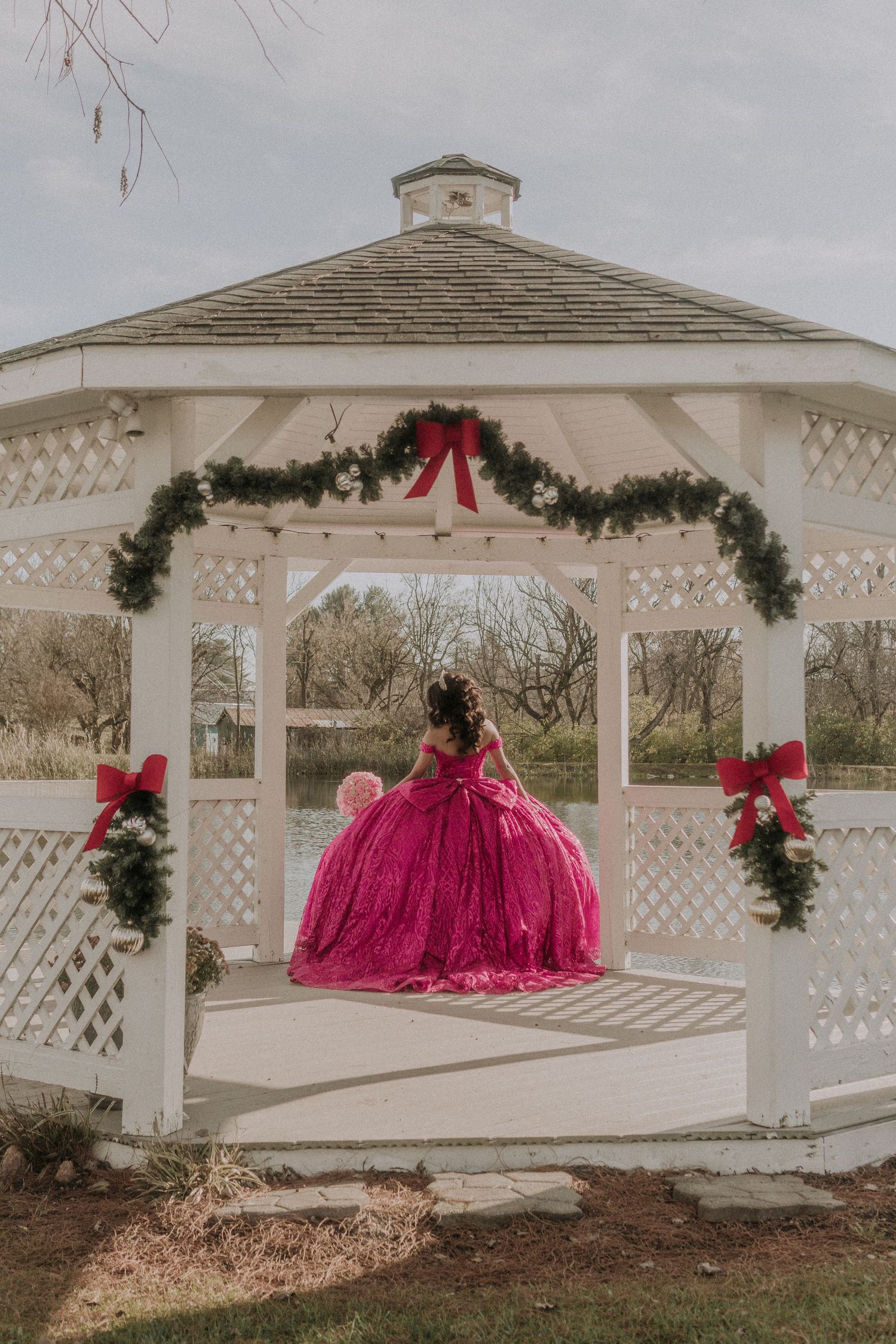 White gazebo decorated with holiday greenery and red bows featuring a girl in a magenta ballgown.